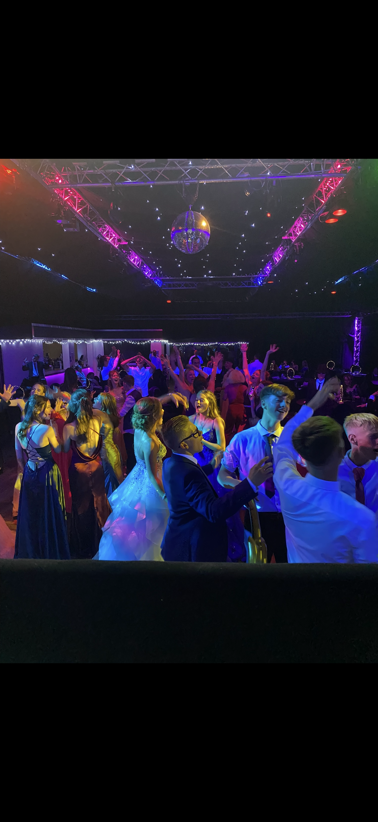 A lively dance party with people in formal dresses and suits dancing and enjoying music under colorful lighting and a disco ball.