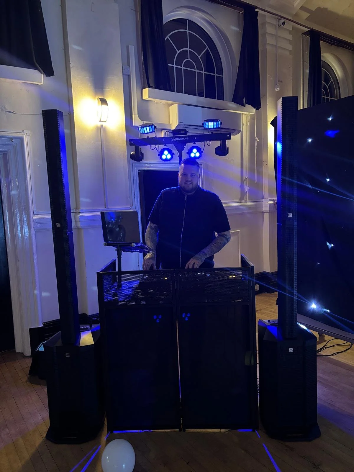 DJ standing at a DJ booth with speakers and lighting equipment in a decorated indoor venue with arched windows and blue lighting.