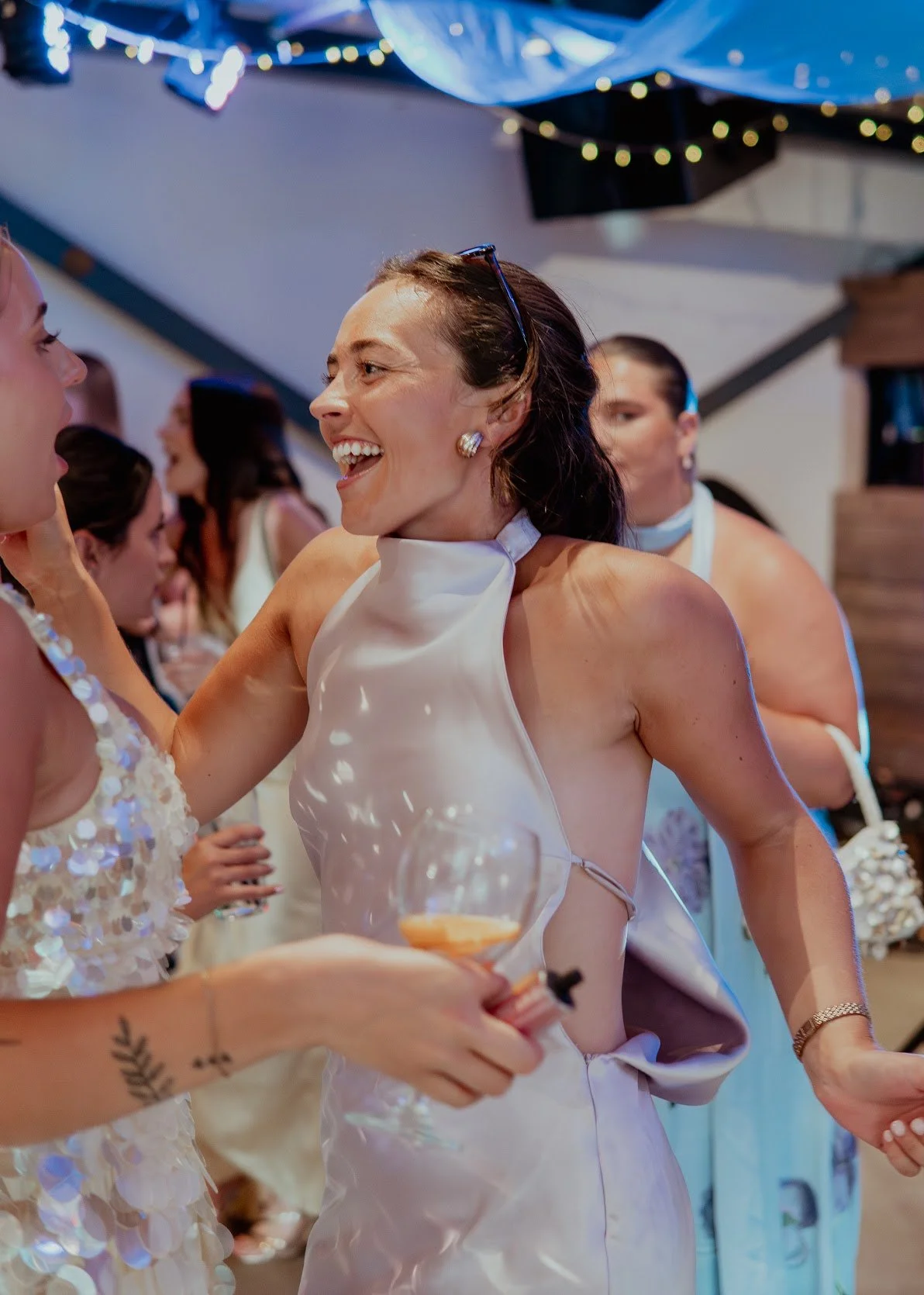 Two women at a wedding reception, smiling and talking, with other guests in the background. One woman is wearing a white dress and holding a drink.