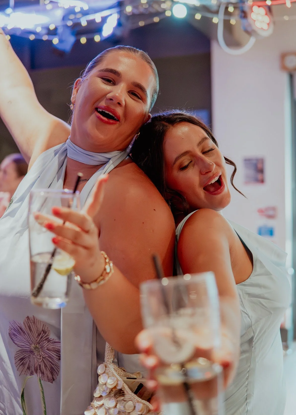 Two women are enjoying a celebration at a bar or restaurant, smiling and holding drinks, with colorful string lights and decor in the background.