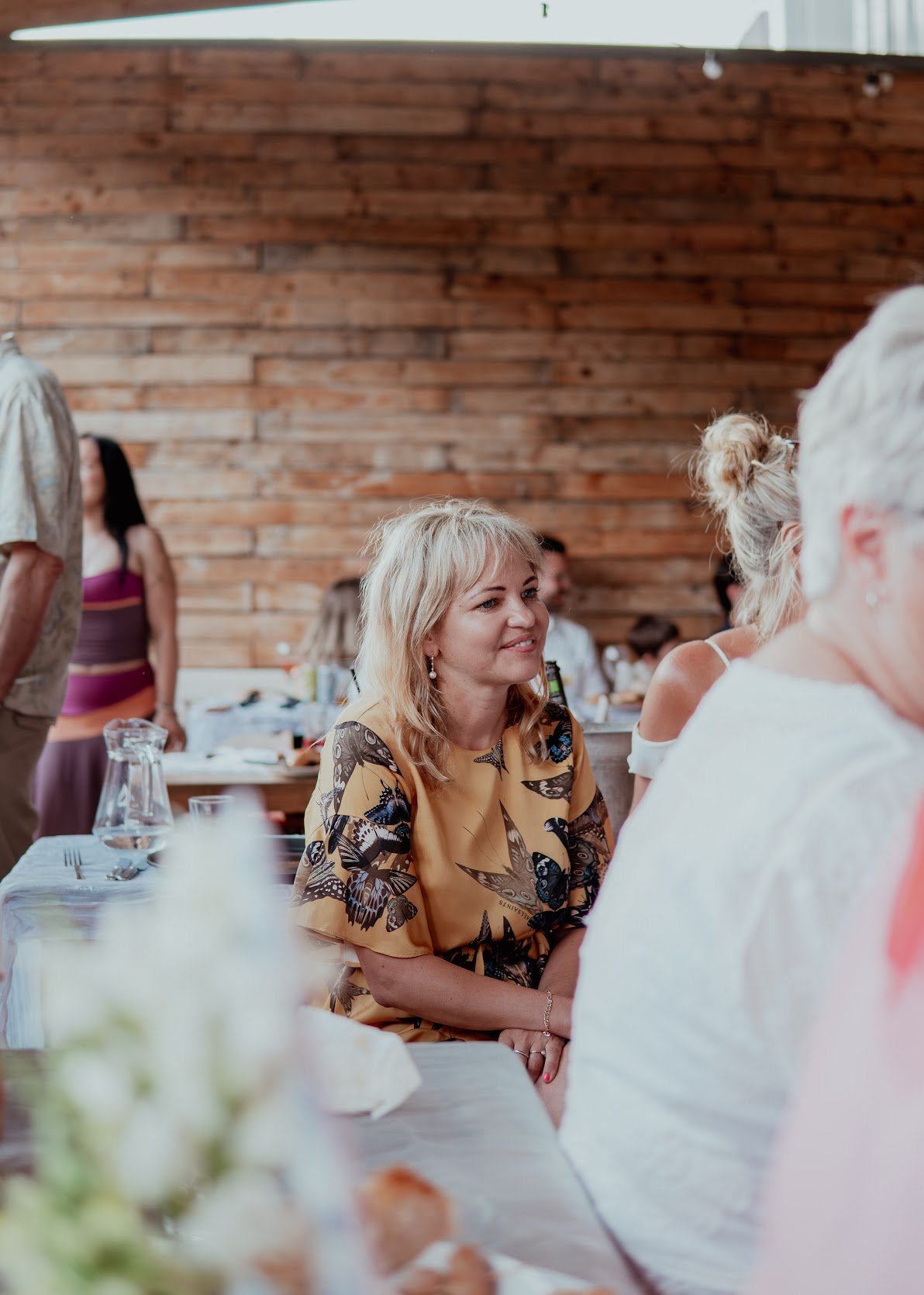 A woman with blonde hair wearing a butterfly-patterned dress smiling at a social gathering inside a venue with wooden walls, surrounded by other people.