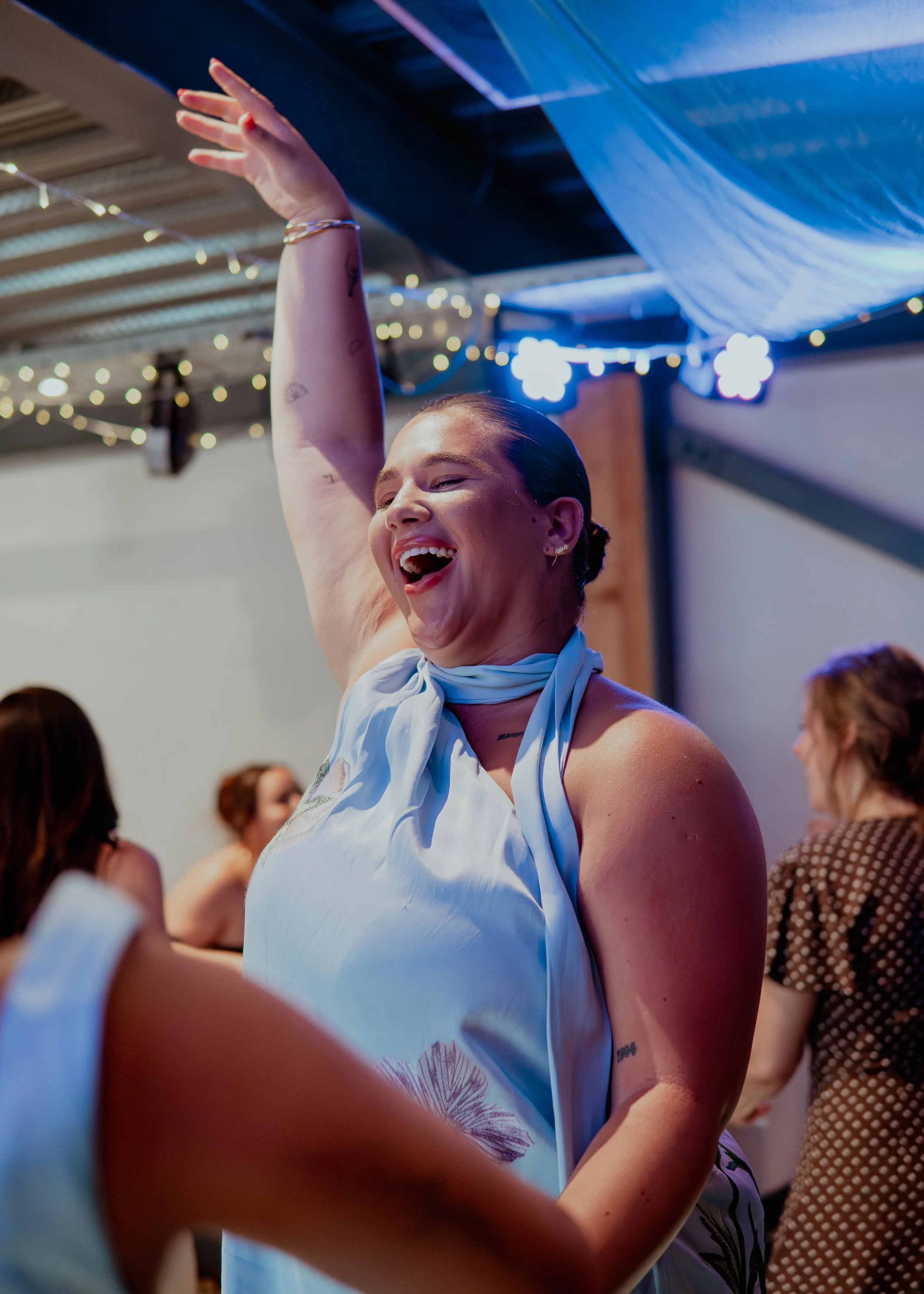 A woman with a joyful expression, wearing a white dress and a light blue scarf, raising her hand in the air at a festive indoor event decorated with string lights and blue drapes.