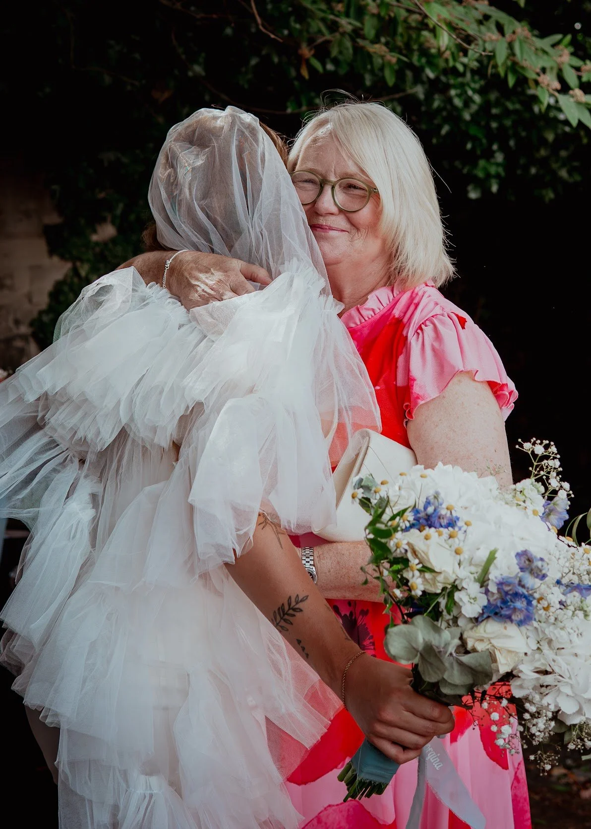 A woman with blonde hair, glasses, and a pink dress hugs another woman dressed as a bride, who is wearing a white wedding gown and veil. The bride is holding a bouquet of white and purple flowers, and the scene is outdoors with green foliage in the b