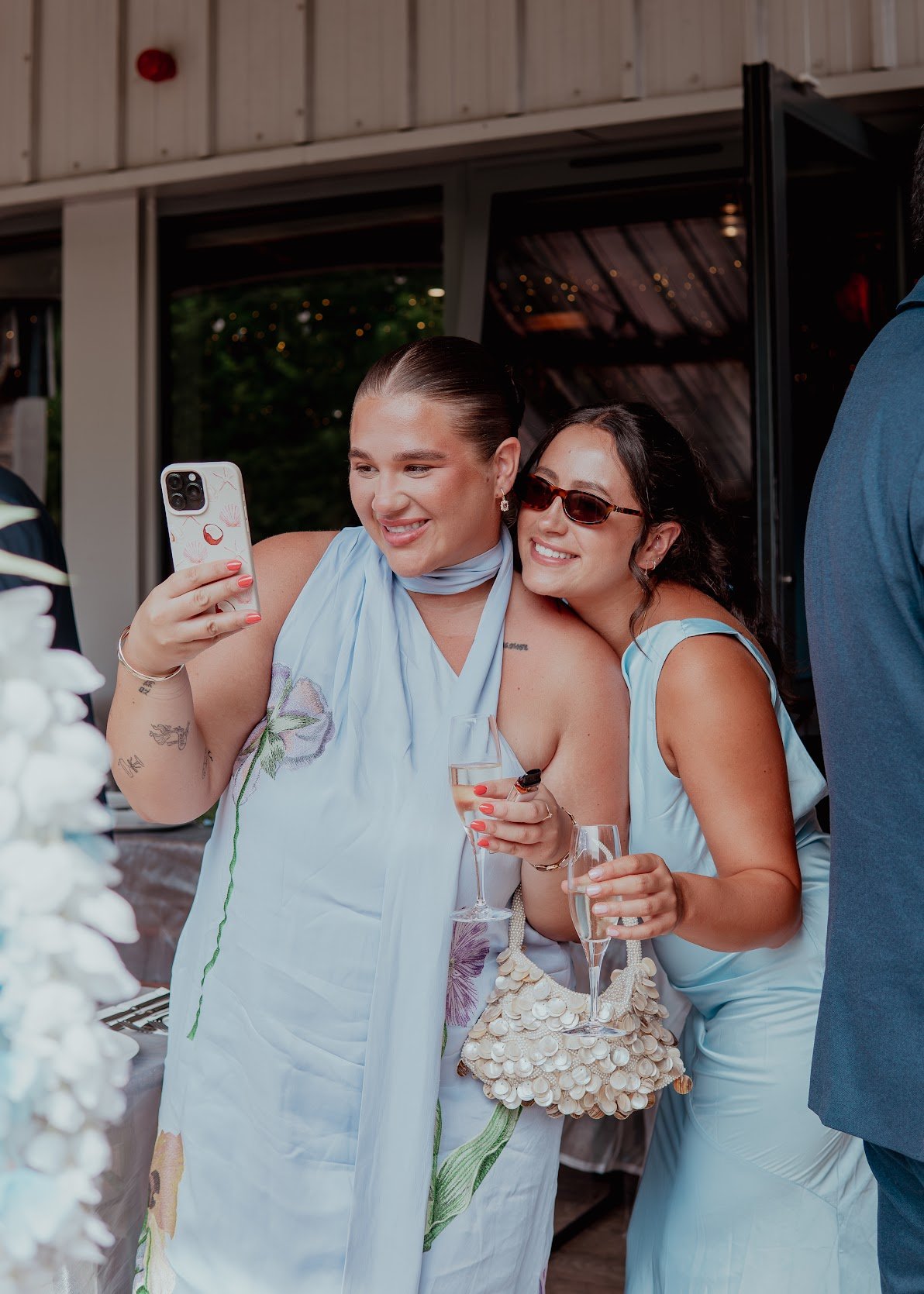 Two women at a celebration taking a selfie, one holding a phone and the other holding a glass of champagne, both smiling, wearing light blue dresses, one wearing sunglasses, inside a decorated venue.