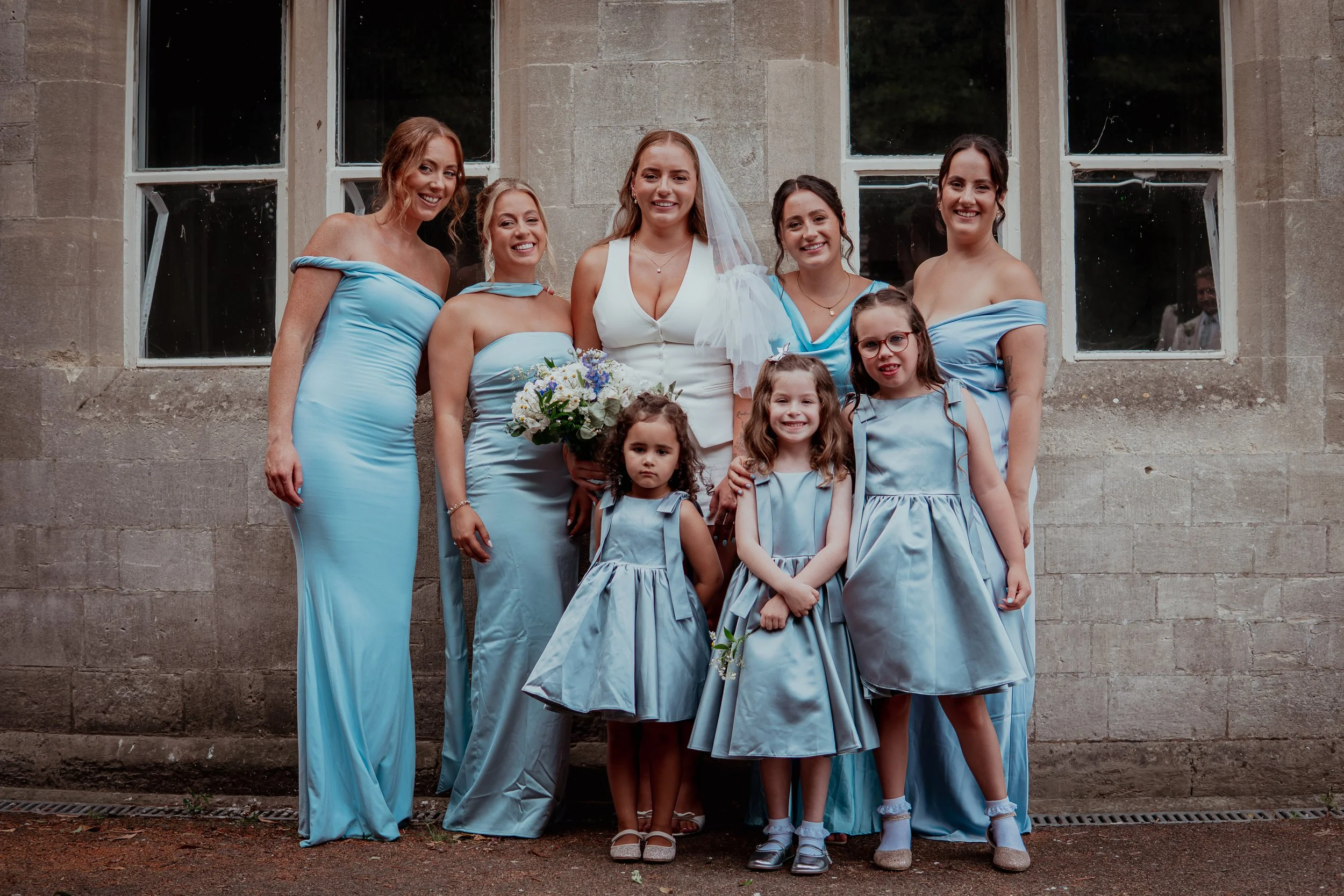 A group of seven women, including a bride in a white wedding dress with veil and bouquet, and six bridesmaids in matching light blue bridesmaid dresses, standing outdoors in front of a stone building with large windows, smiling for a photo.