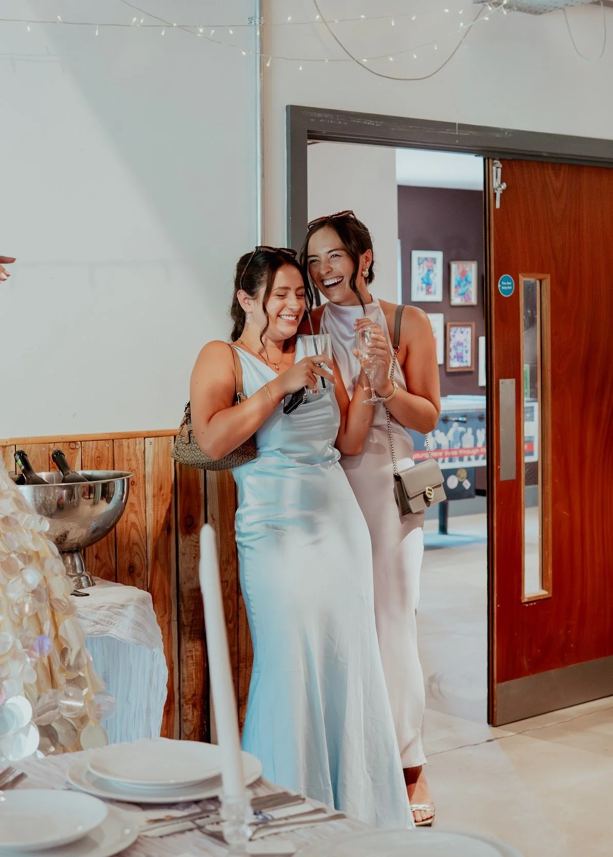 Two women dressed in white, hugging and smiling, holding champagne glasses at a celebration or wedding reception. One has long dark hair and the other has short dark hair. They are standing near a table with a white candle, some plates, and bottles o