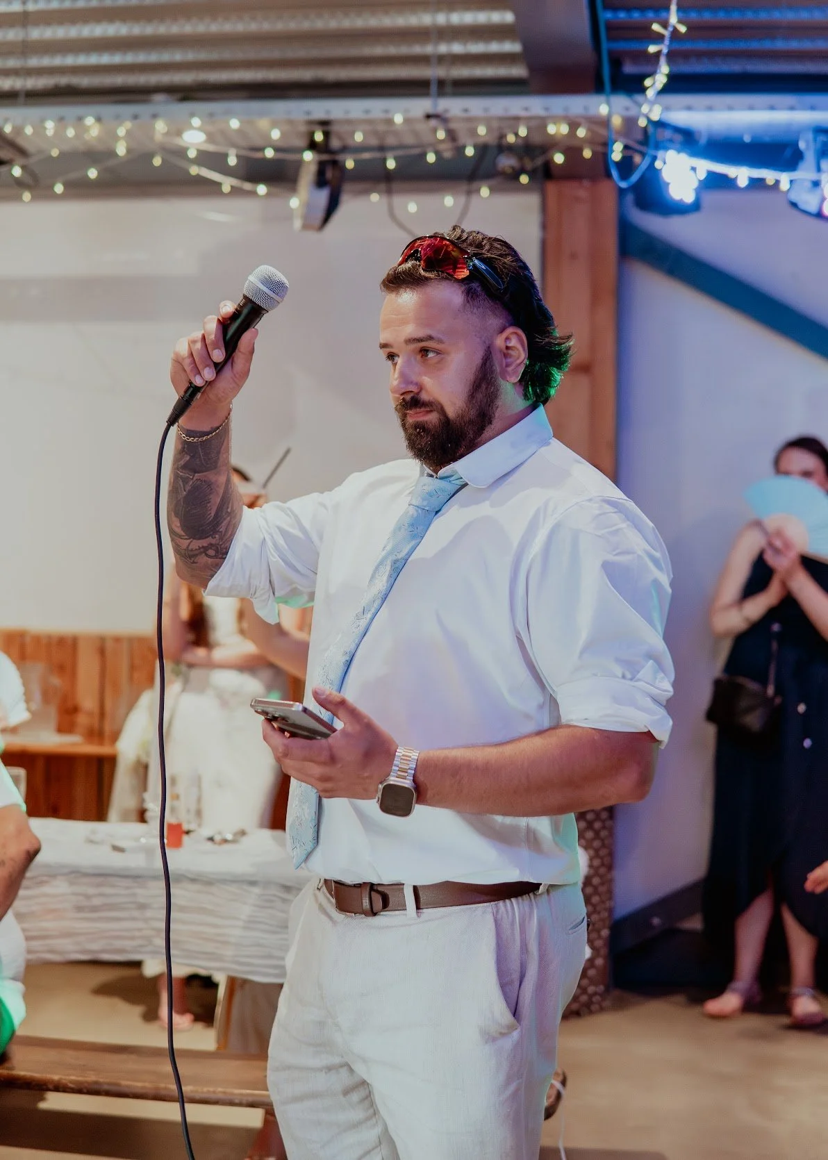 Man with beard and tattoos holding a microphone and a smartphone at a social gathering with string lights and other people in the background.