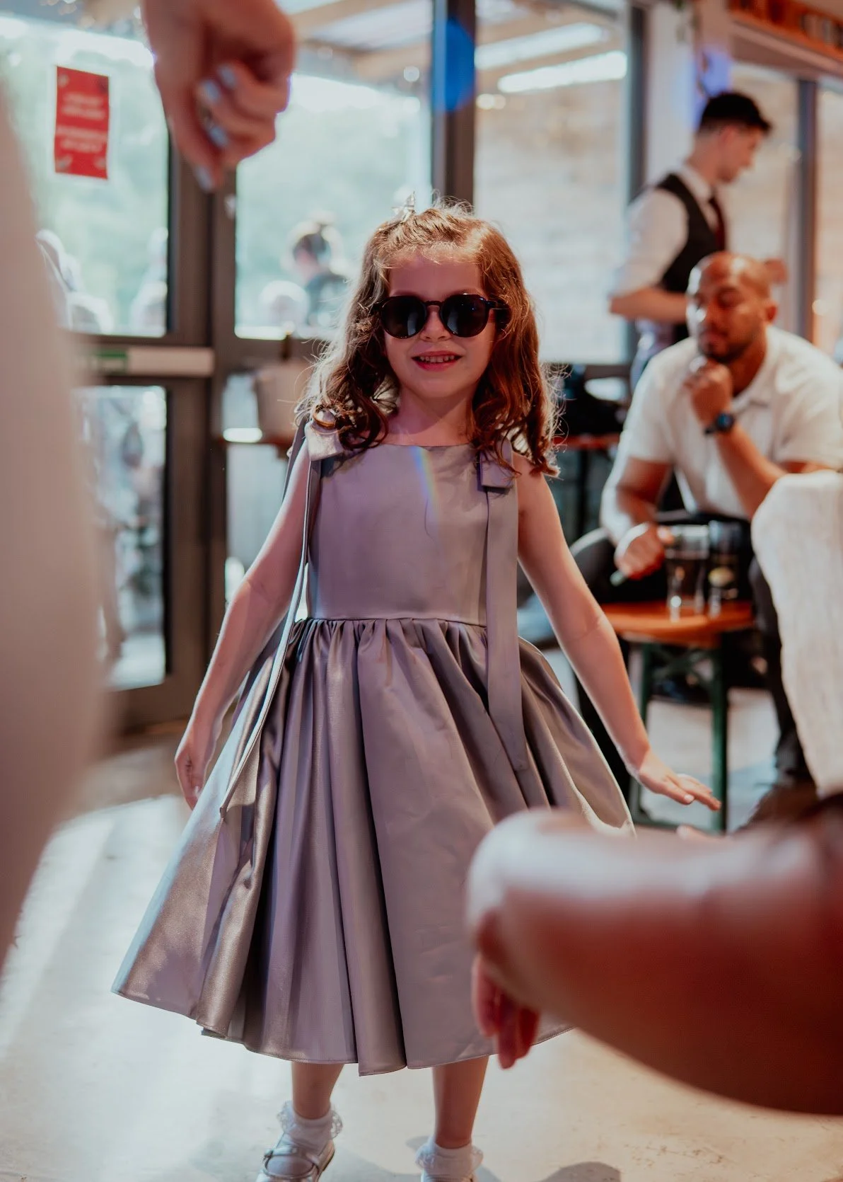 Young girl with curly hair wearing sunglasses and a shiny, plum-colored dress, smiling and walking in a restaurant or cafe. Background shows other diners and waitstaff.