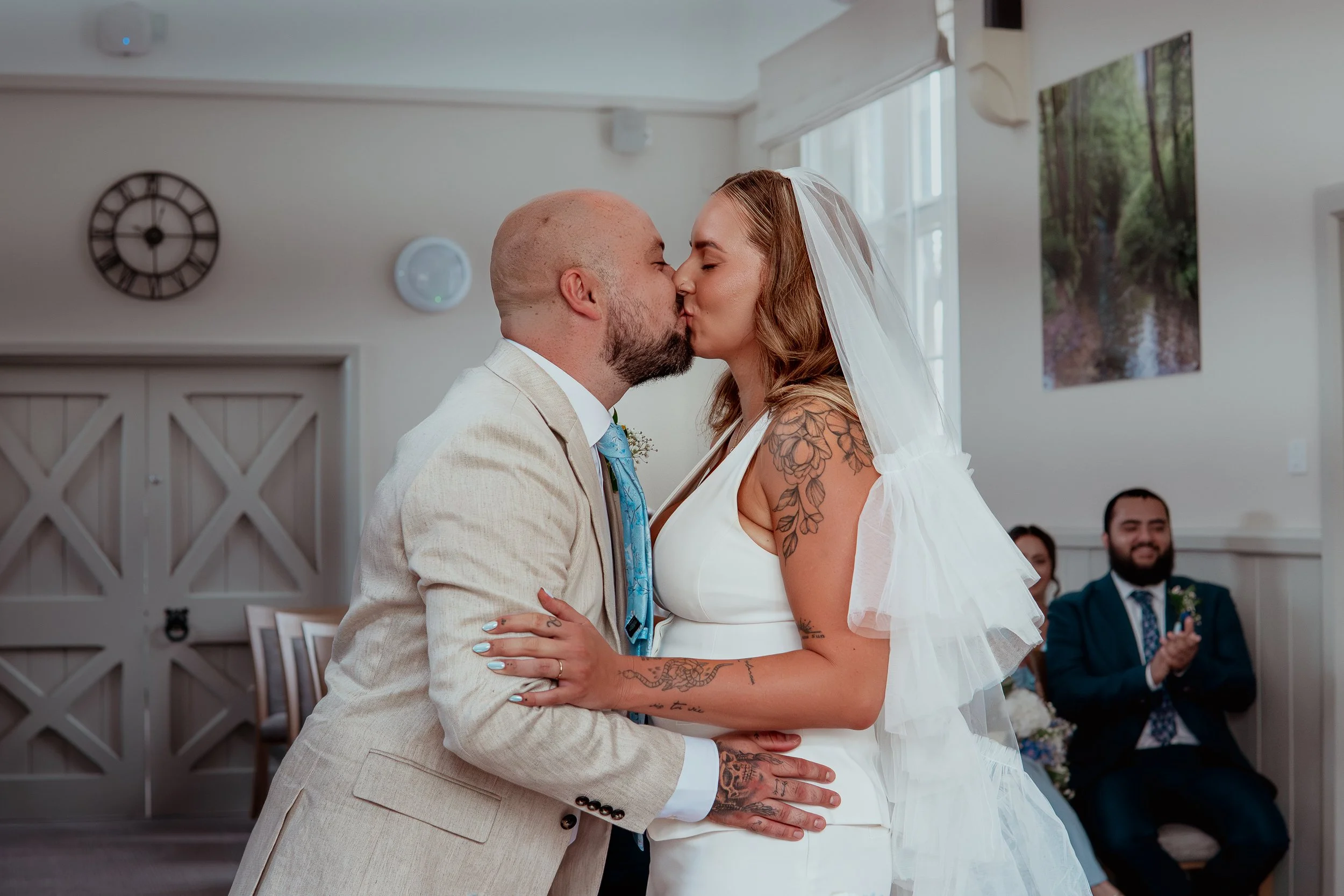 A couple in wedding attire sharing a kiss during their wedding ceremony inside a decorated room with seated guests and artwork on the walls.