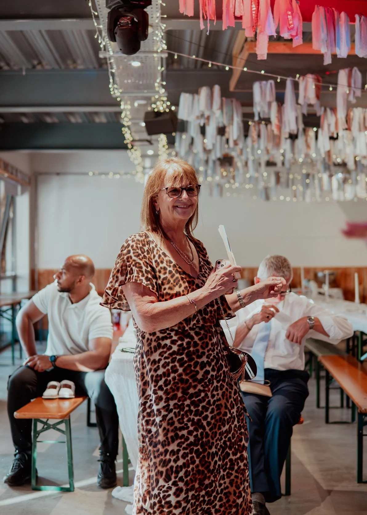 A woman in a leopard print dress and sunglasses smiling while holding a phone in a festive indoor setting, with hanging pink and white decorations overhead, and two men seated in the background.