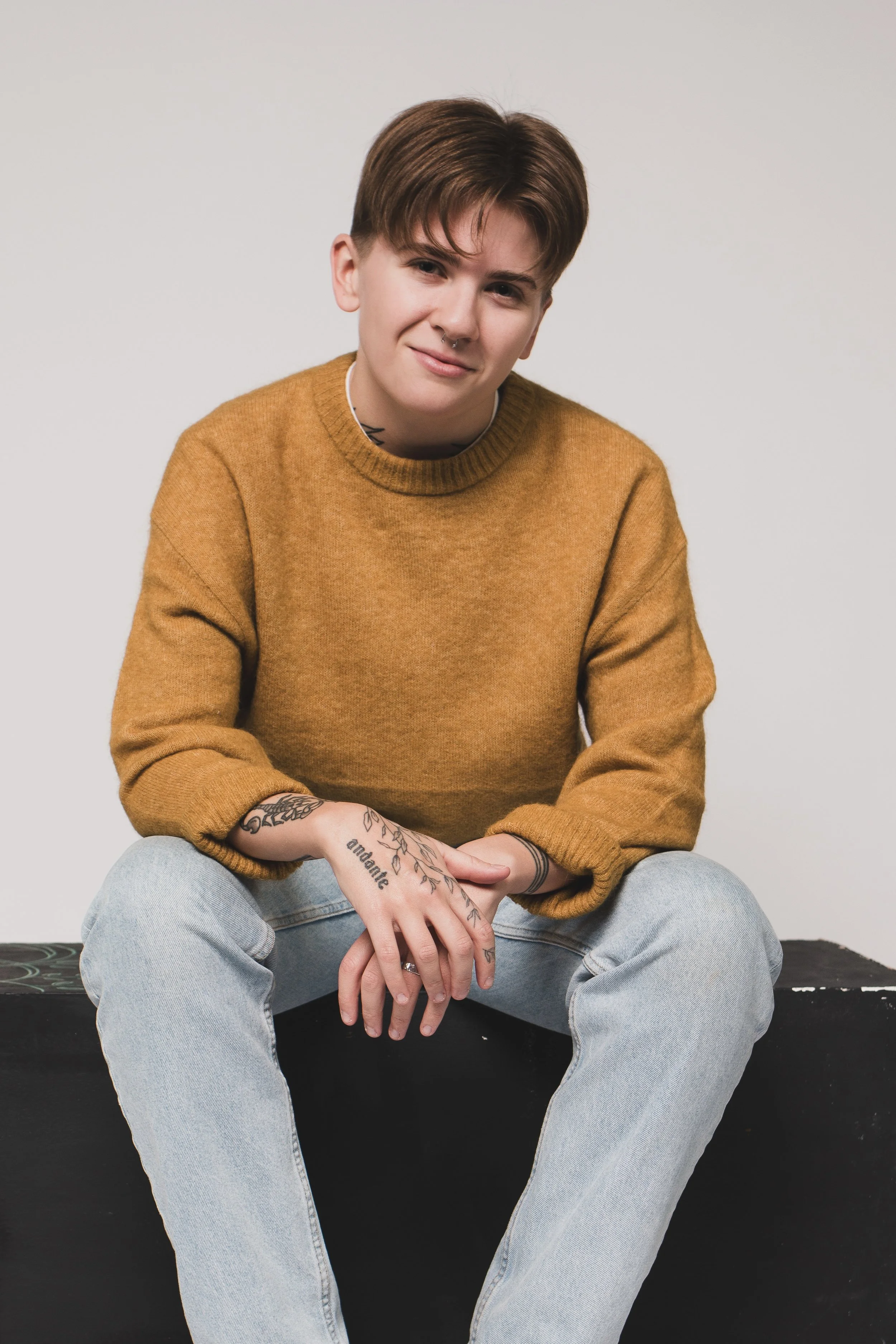Portrait of a young person with short brown hair, wearing a mustard sweater and light jeans, sitting on a black platform against a plain light gray background.