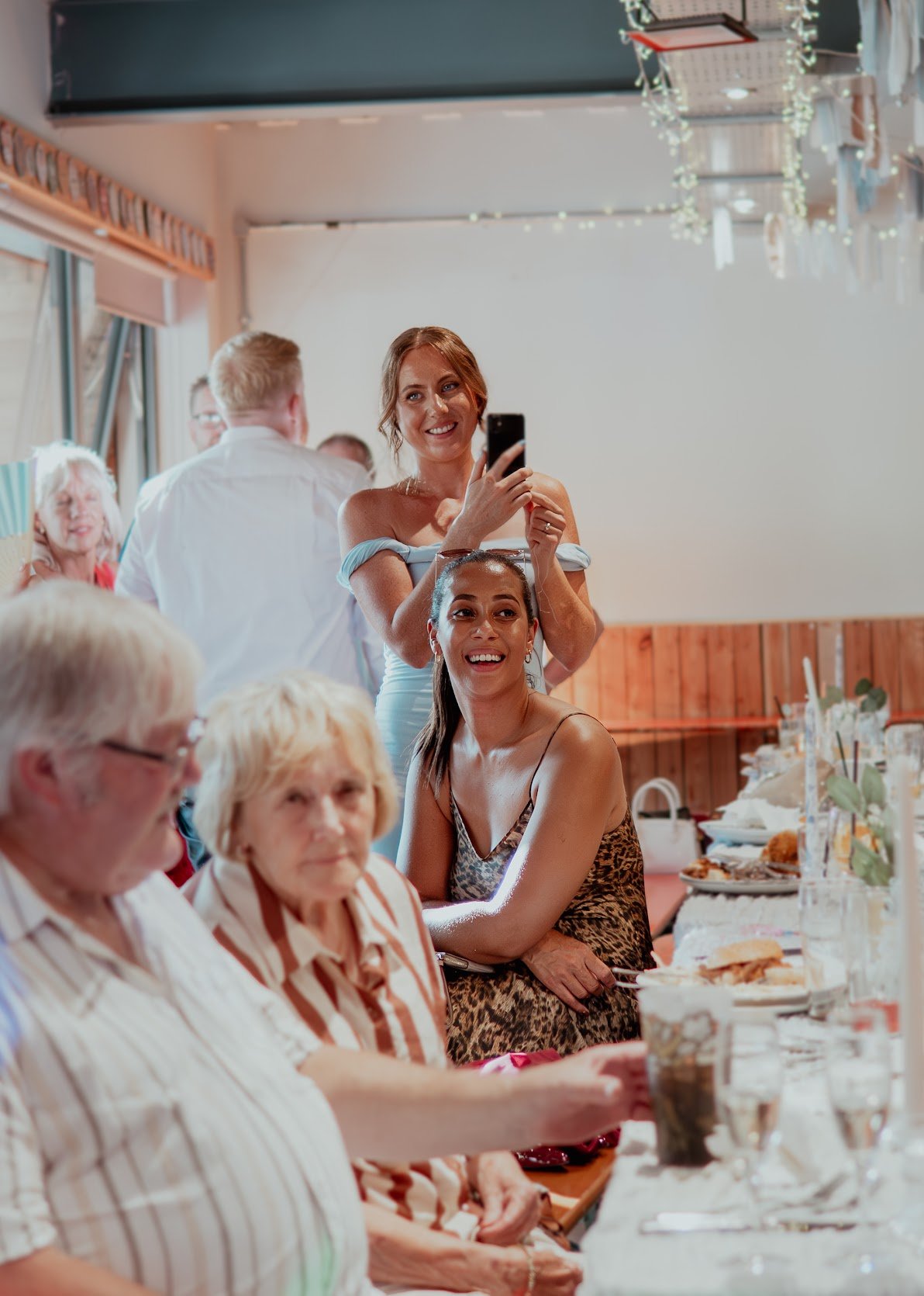 A woman standing and taking a photo with her phone while others sit at a table during a social gathering or celebration in a decorated indoor setting.