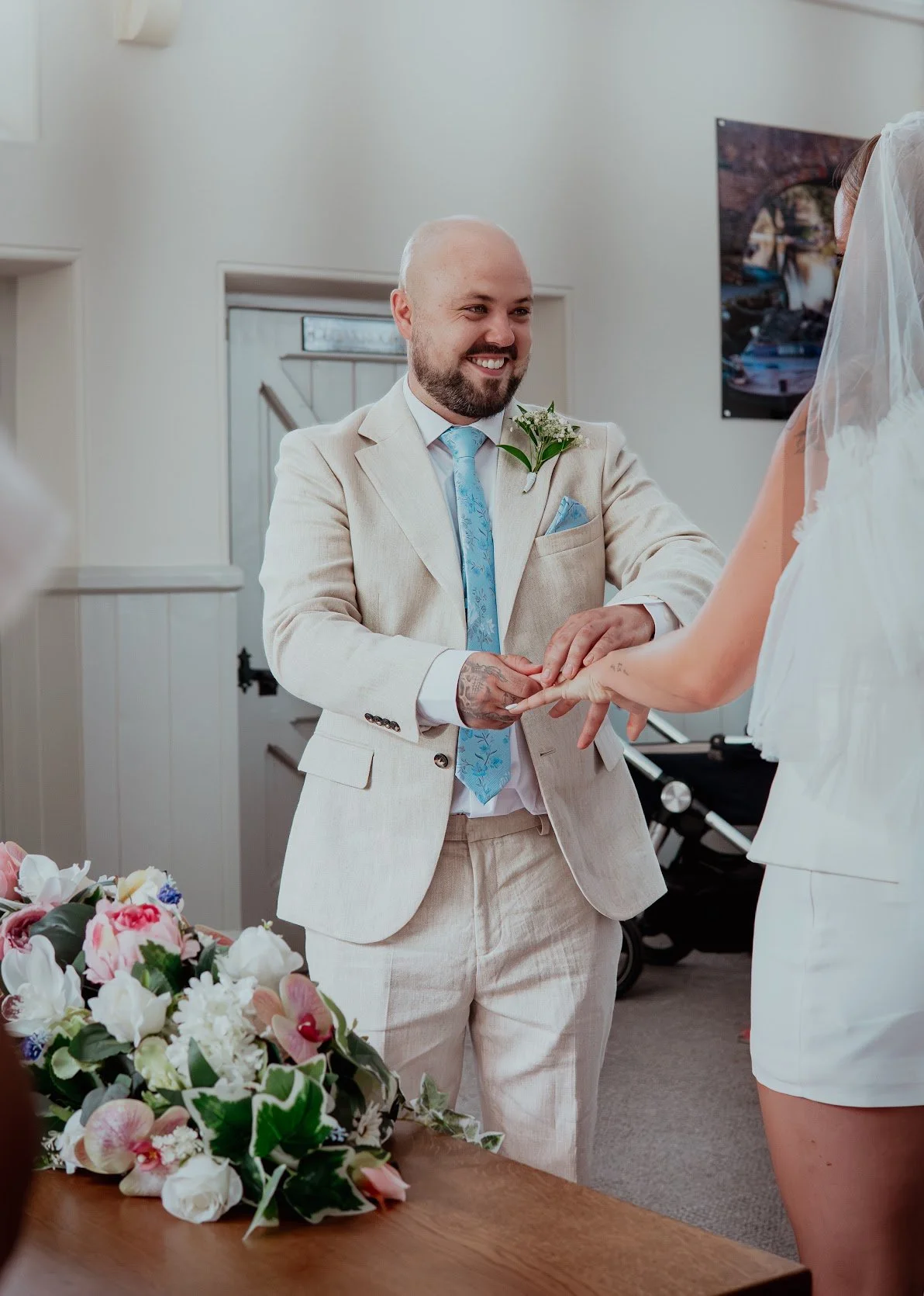A groom in a cream suit exchanging rings with a bride in a white dress and veil during a wedding ceremony indoors. There is a floral arrangement on a wooden table in the foreground.