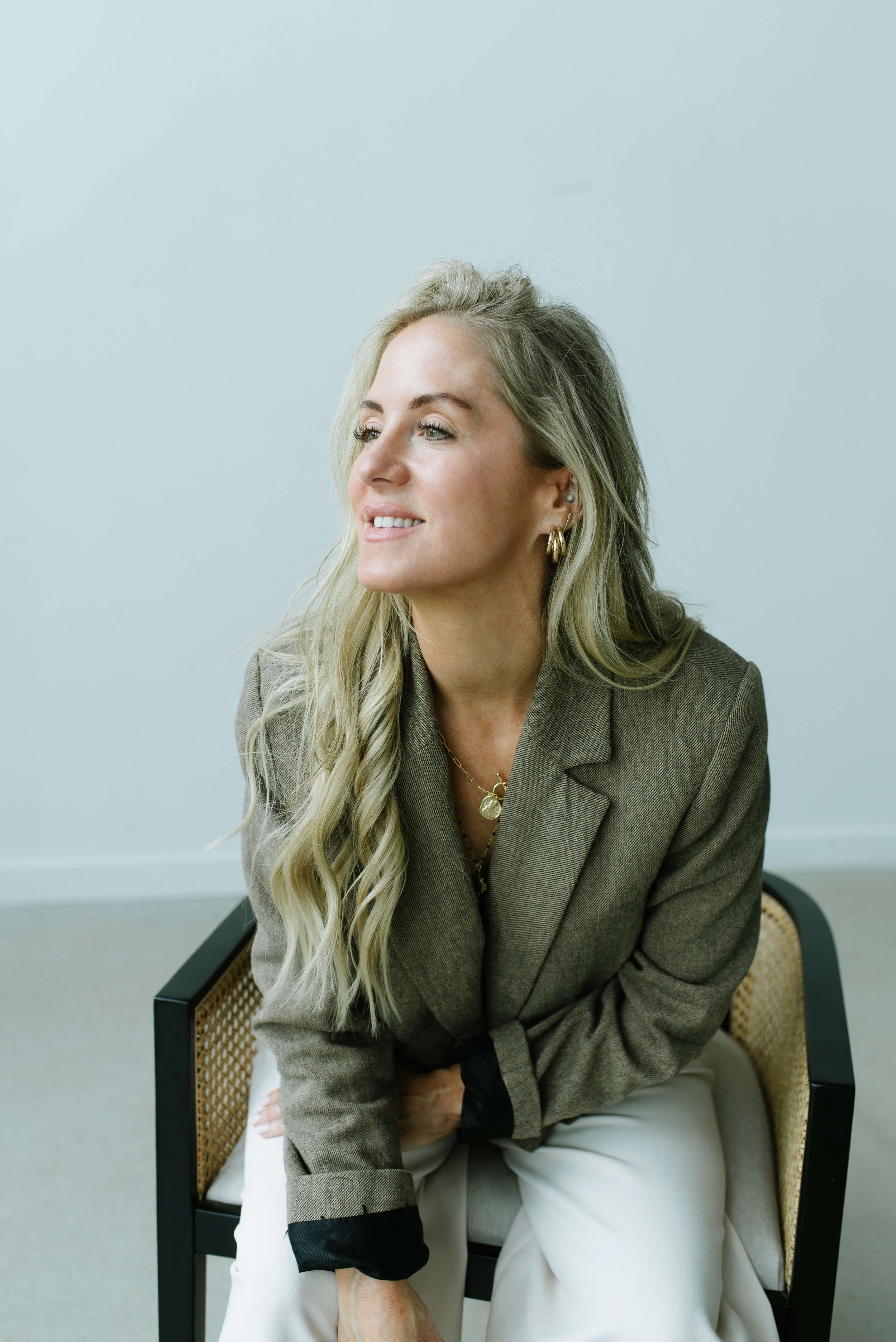 Dani Lynch, a white woman with long, wavy blonde hair wearing a gray blazer and gold jewelry sits on a chair in front of a plain, light-colored wall.