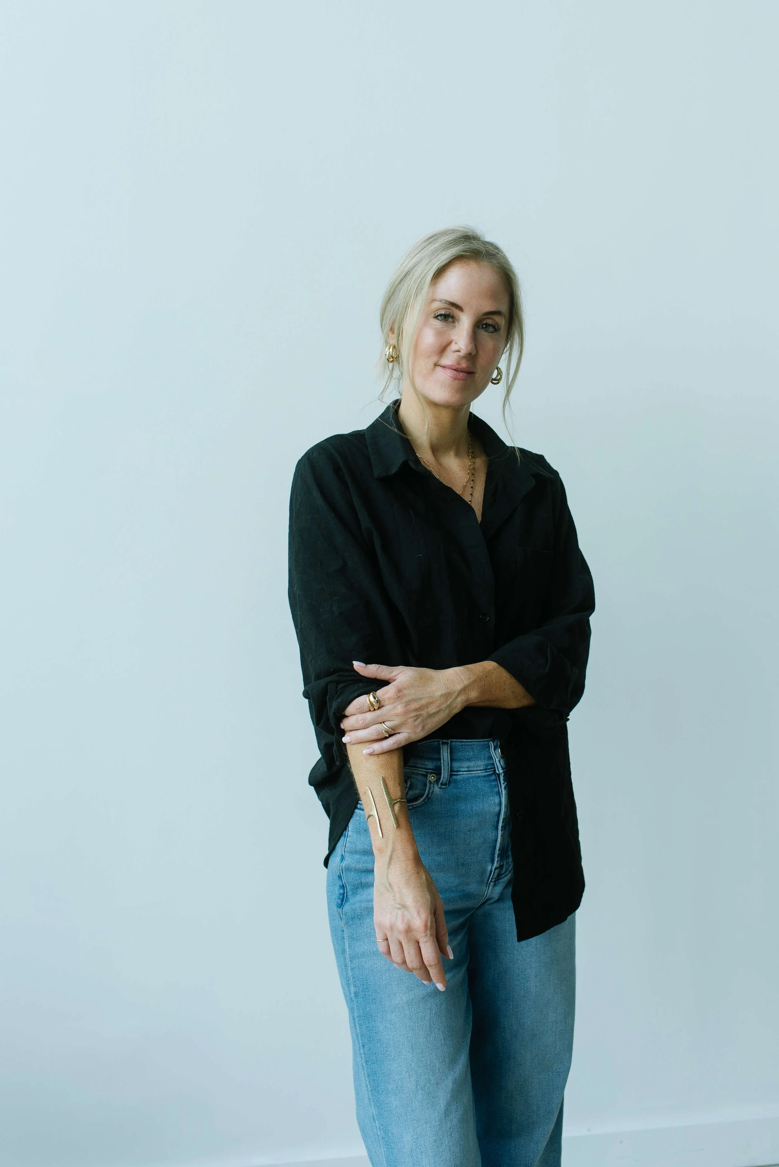 Dani Lynch, a white woman with blonde hair styled in a loose side bun wears a black button-up shirt and blue jeans, and stands against a light blue background.