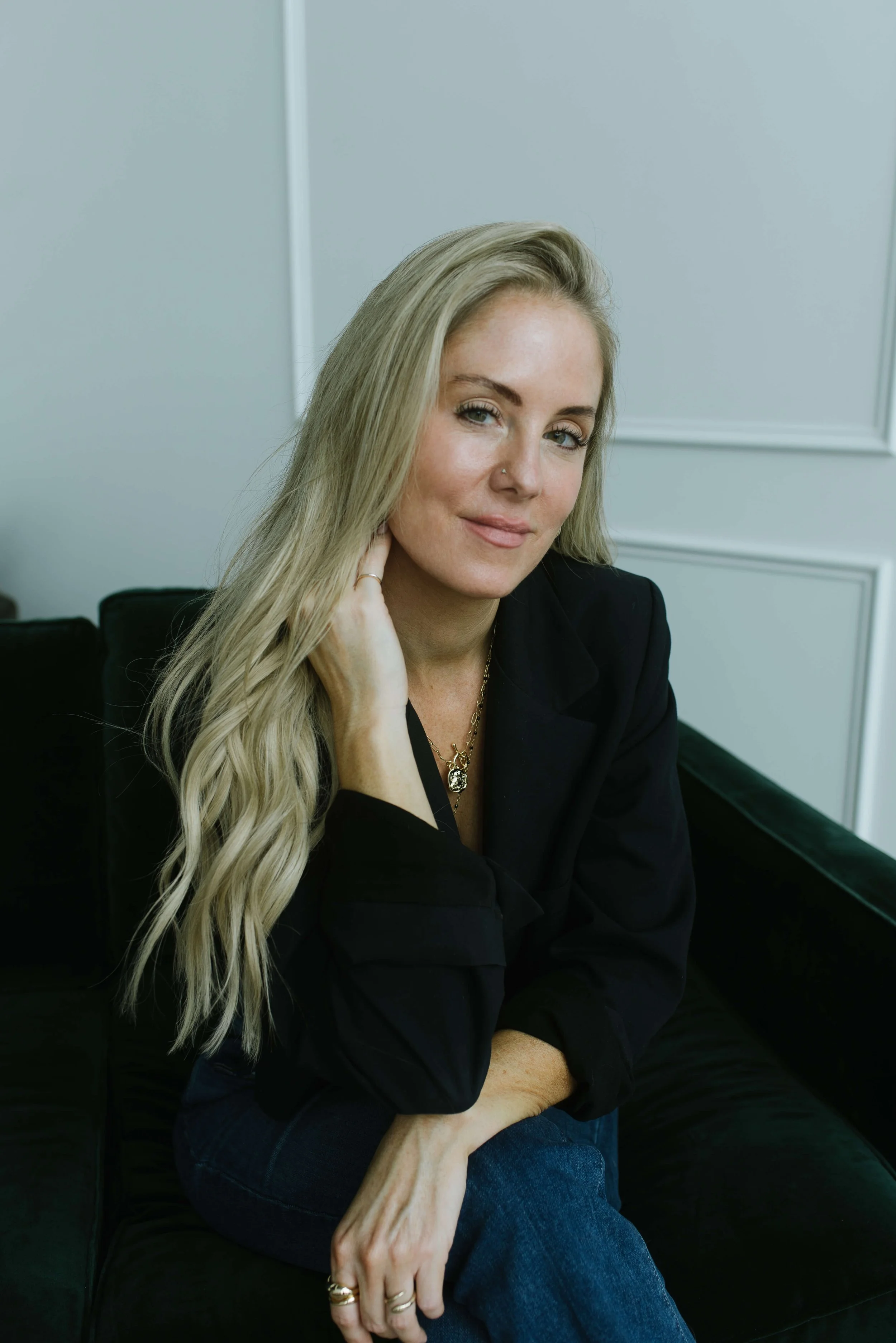 Dani Lynch, a white woman with long blonde hair, is wearing a black blazer, sitting on a black couch with a light gray wall behind her, smiling softly and looking at the camera.