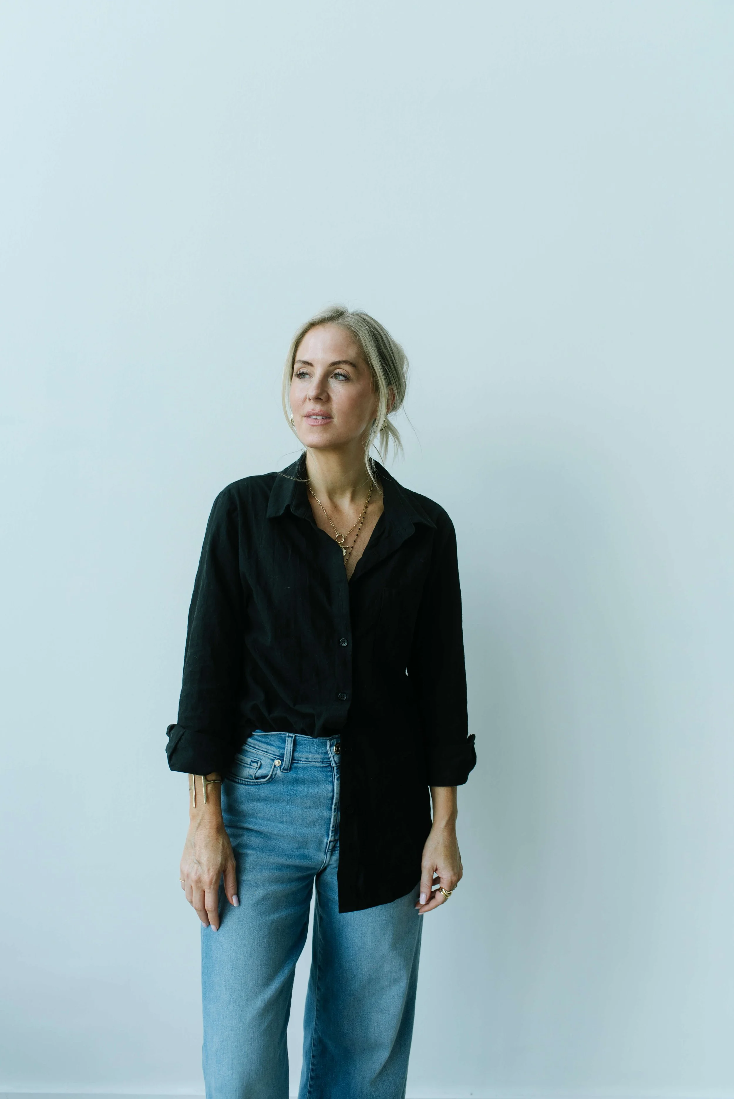 Dani Lynch, a white woman with blonde hair styled in a loose bun, wears a black button-up shirt and light blue jeans, stands against a light-colored wall, and is looking slightly to her right with a neutral expression.
