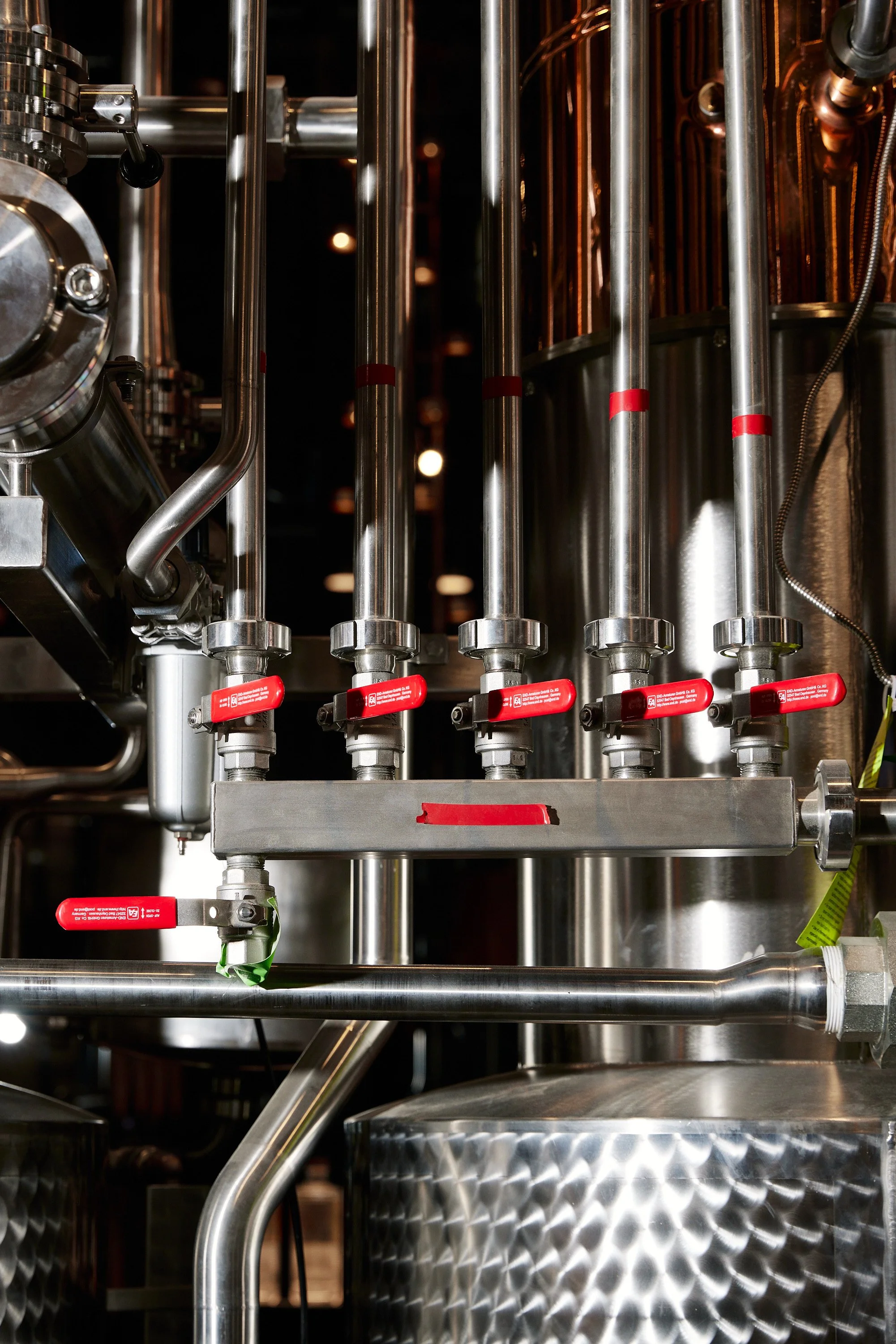 Close-up of stainless steel pipes and valves with red handles in a brewery or industrial setting.