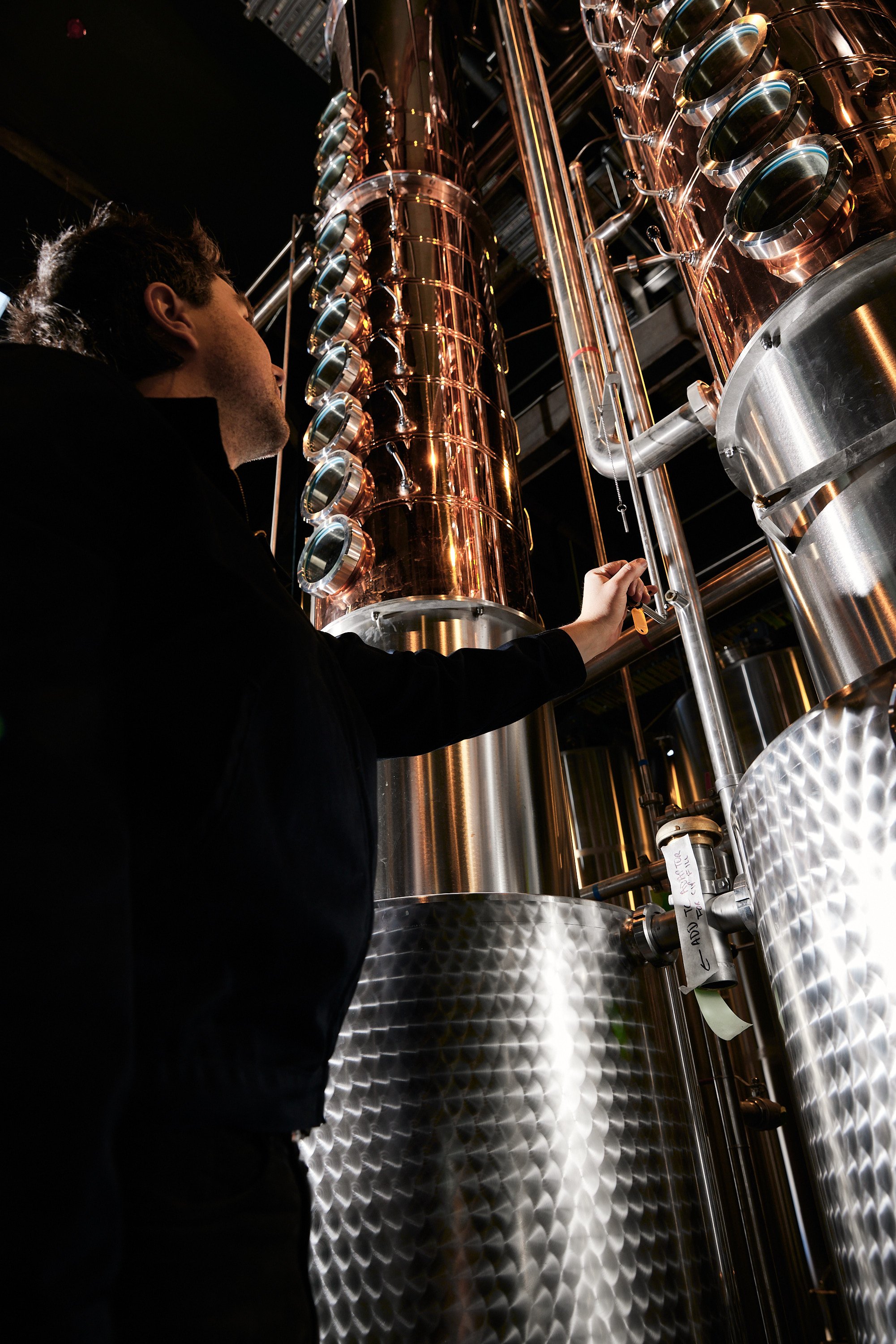 A person inspecting or adjusting a complex industrial machine or distillation apparatus made of shiny metal and copper pipes in a laboratory or factory setting.