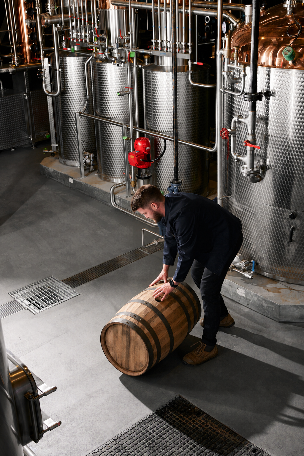 A man in a brewery inspecting a wooden barrel surrounded by large stainless steel tanks and brewing equipment.