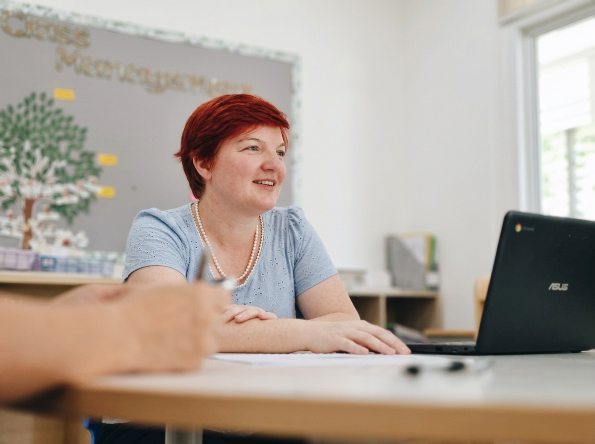 A woman with short red hair sitting at a desk with a laptop and a person in the foreground taking notes, in a classroom or meeting room setting.