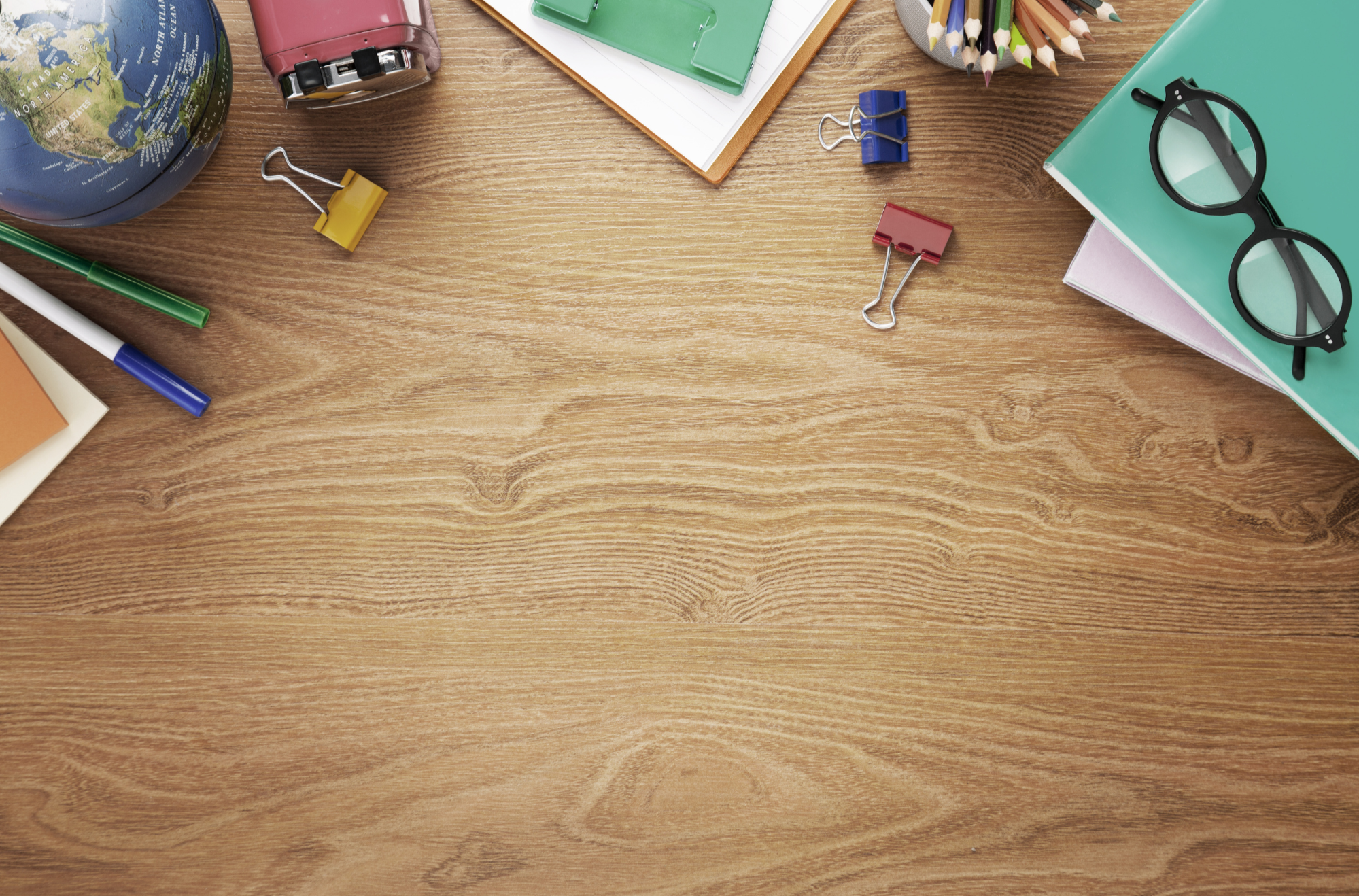 Various school supplies arranged on a wooden desk, including a globe, a pink pencil sharpener, a clipboard with paper, colored pencils in a holder, binder clips, notebooks, and eyeglasses.