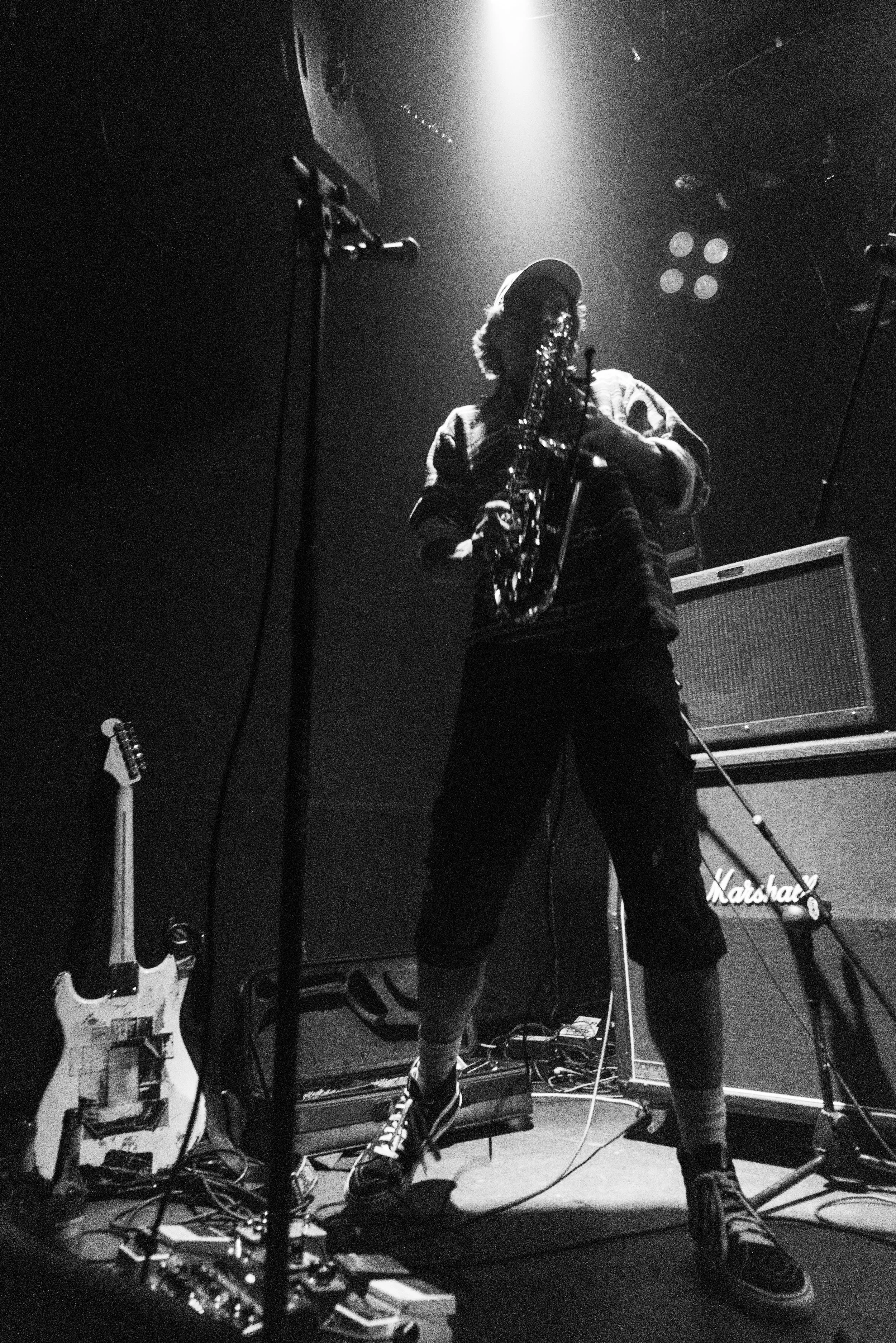 A musician playing a saxophone on a dimly lit stage with spotlights overhead. Equipment including guitars, amplifiers, and microphones are visible around them.