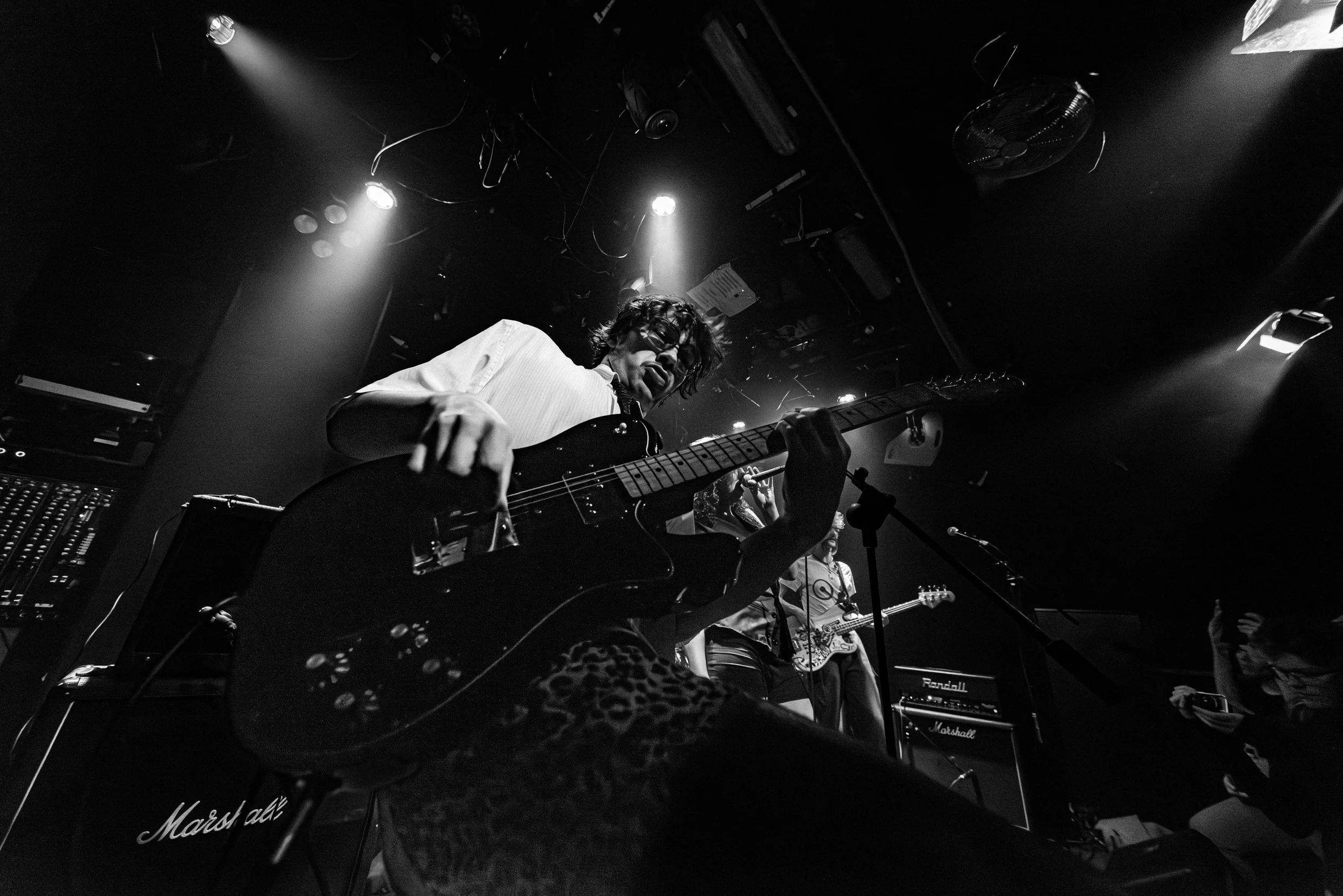 A black and white photo of a musician playing an electric guitar on stage during a live performance. The scene is illuminated by stage lights, with amplifiers and equipment visible in the background.