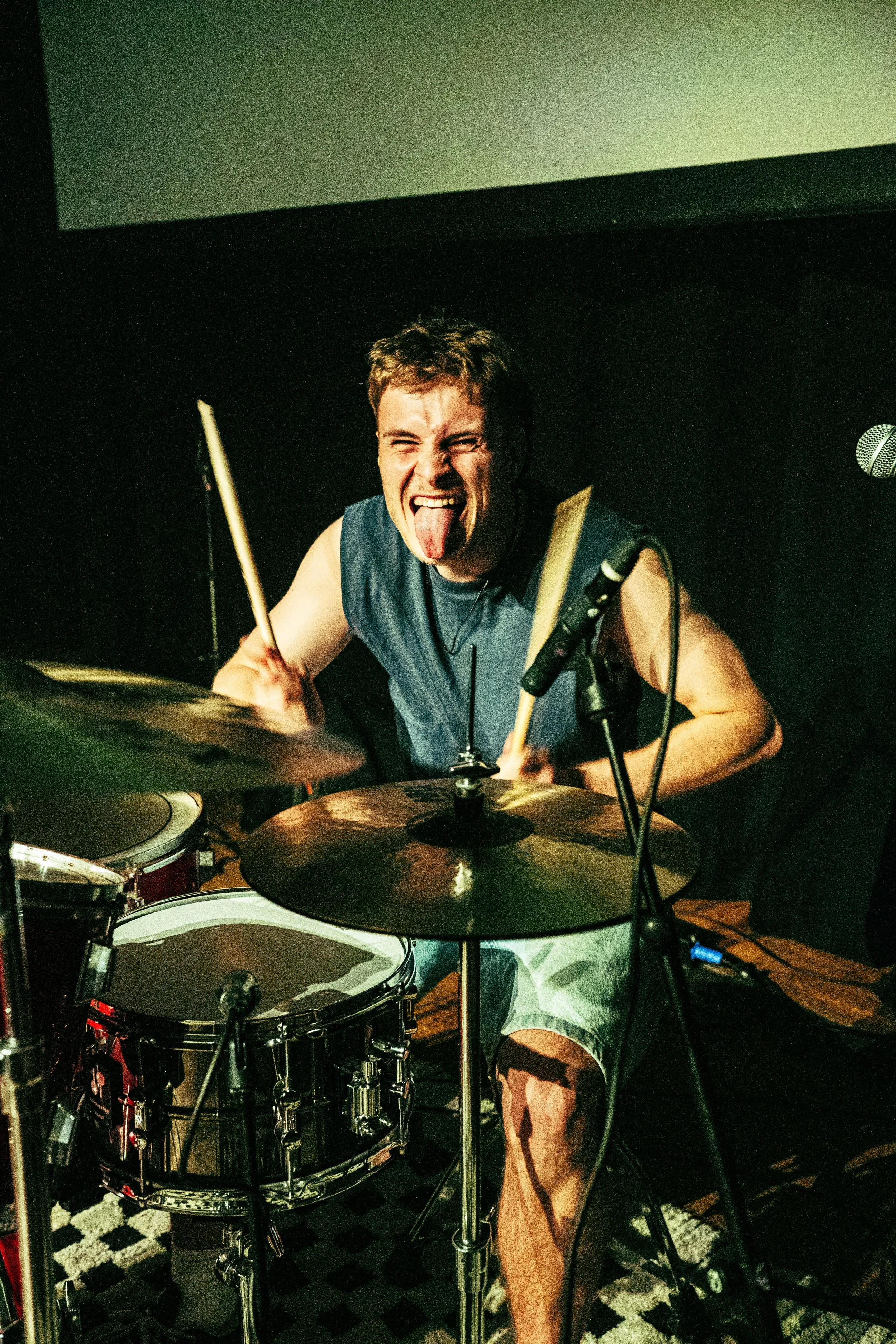 A young man passionate about music is playing drums in a dark room with a black wall and a large blank screen above him. He is sticking out his tongue and has a joyful, energetic expression.