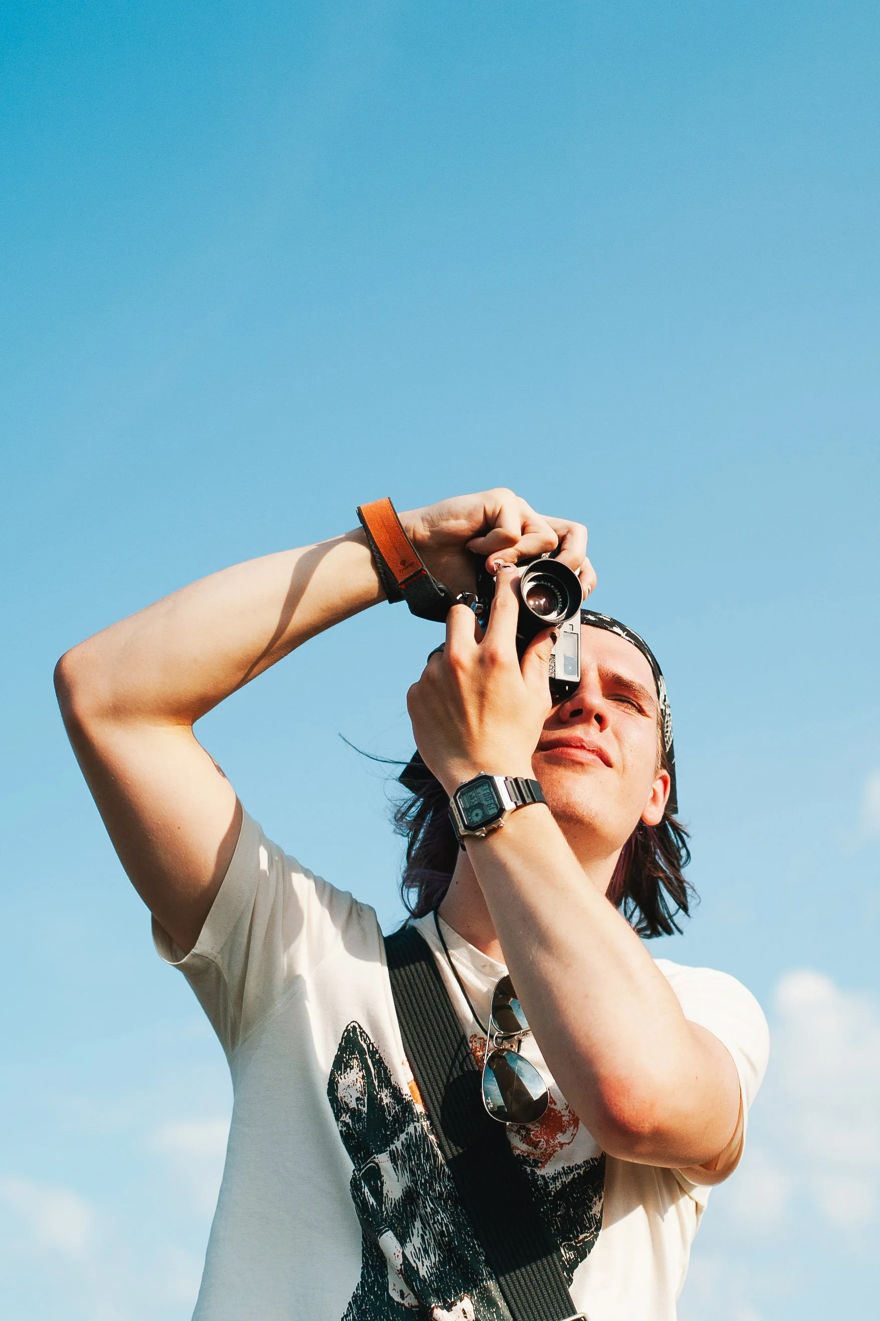 A person with dark hair and a watch on their wrist is holding a leica up to their face while taking a photo, with a bright blue sky in the background.