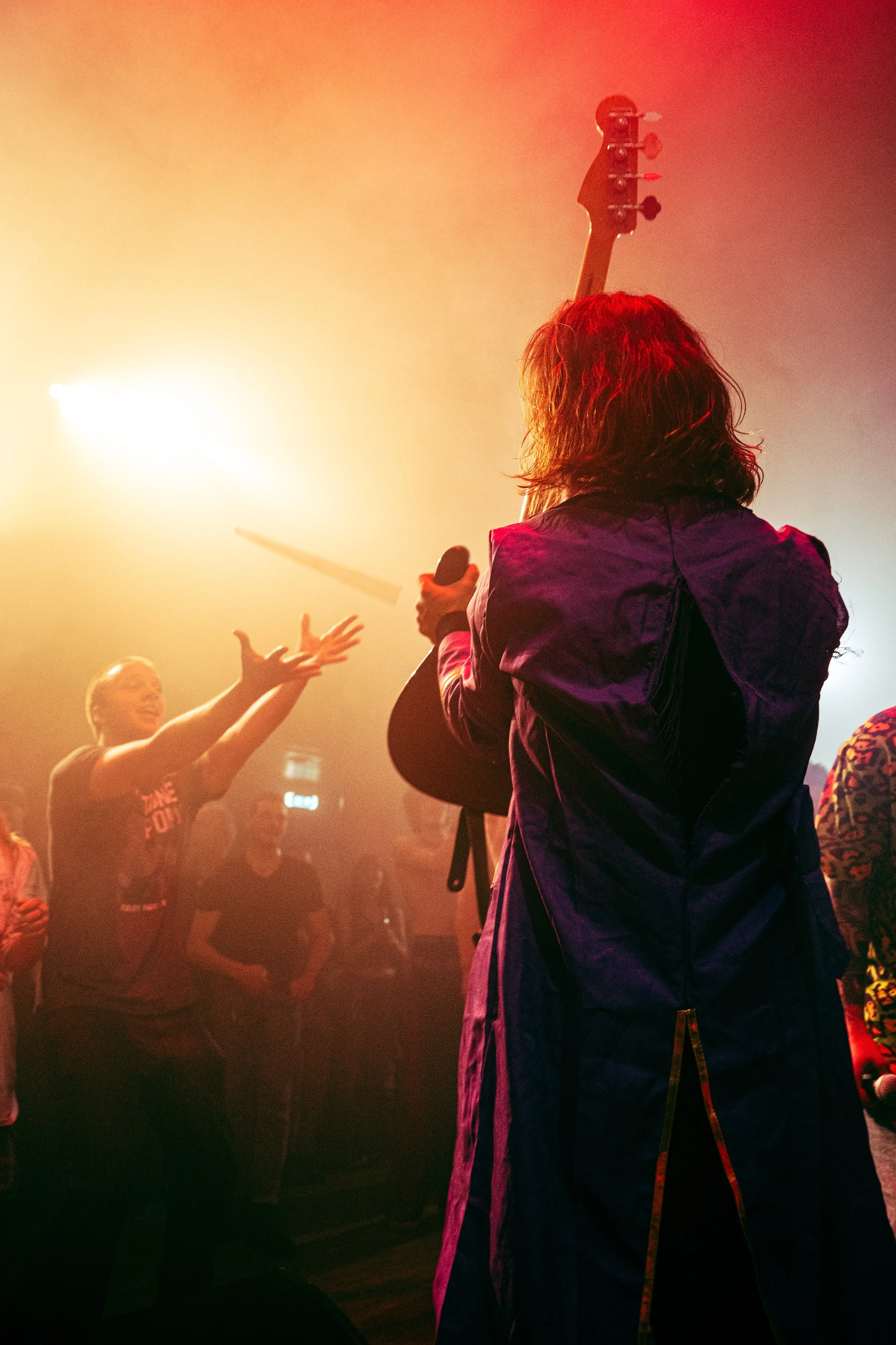 A person with curly hair holding a microphone on stage, back facing the camera, other people reaching out hands towards them, amid colorful lighting and fog.