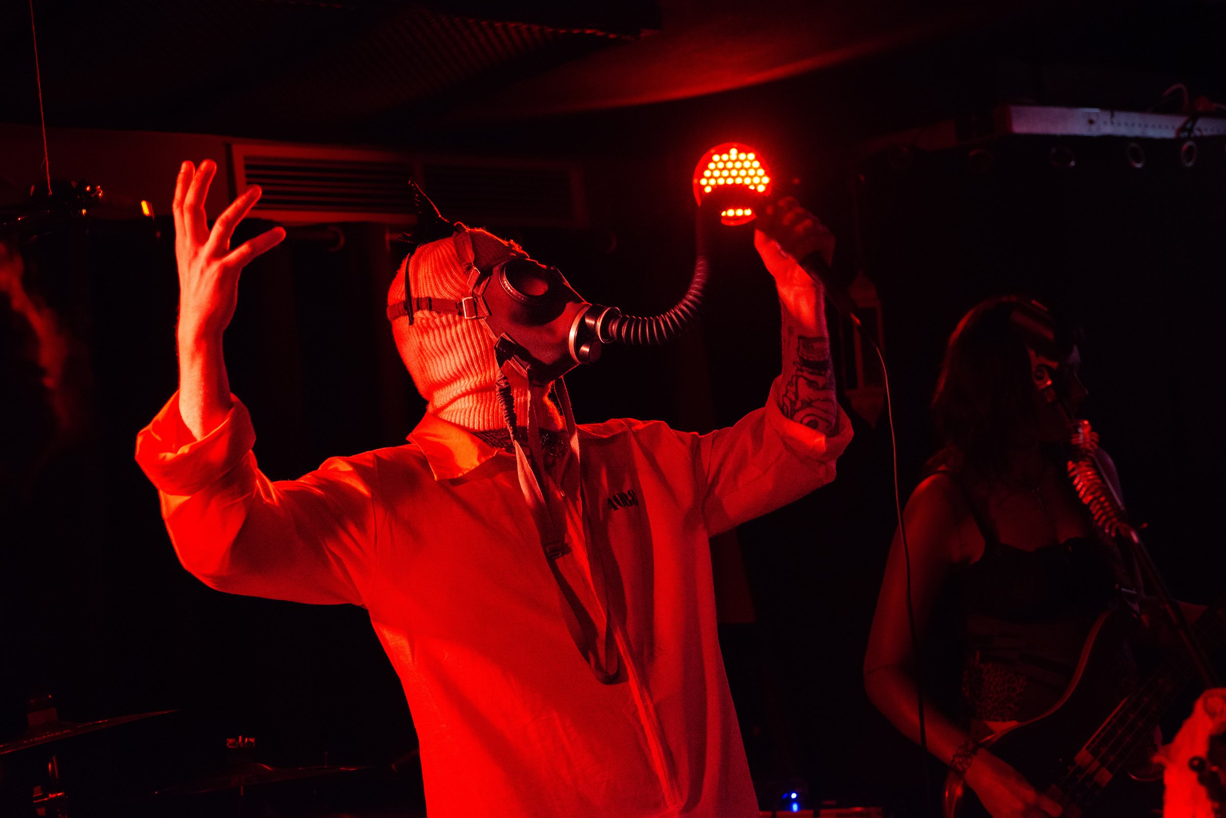 Person wearing a gas mask and hoodie holding a large red LED light in a dark setting, with other people nearby.