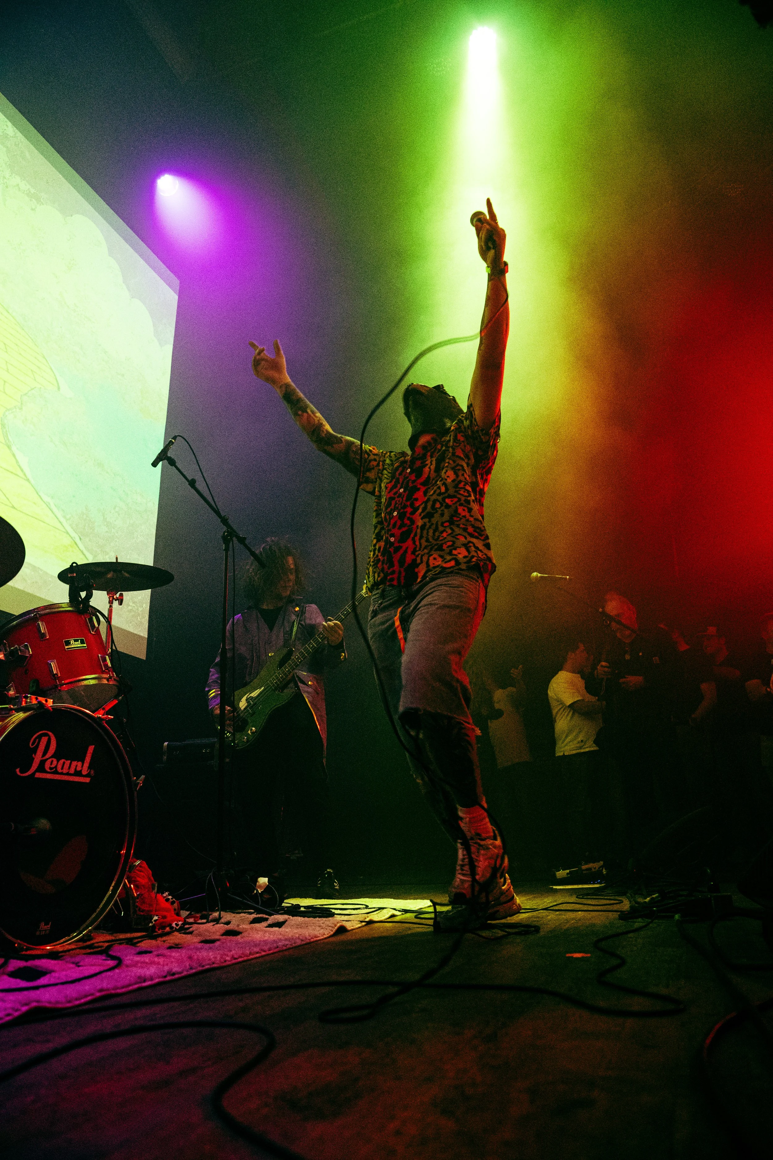 A live music performance on stage with a singer in a patterned shirt raising their arms, colorful stage lights, musicians playing instruments, and a drum set labeled 'Pearl' in the foreground.
