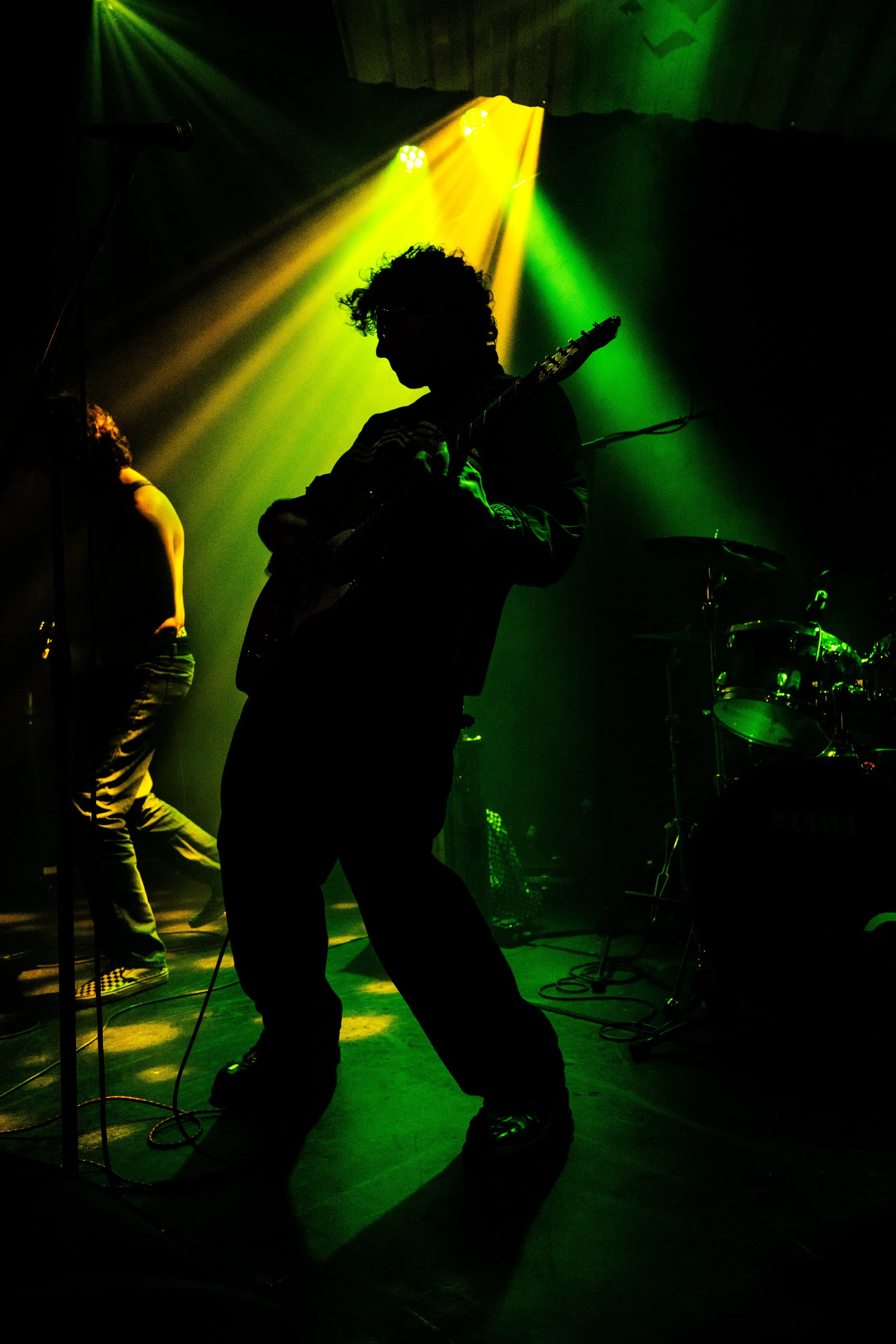 Silhouette of a guitarist on stage with green and yellow stage lights shining down, a microphone stand to the left, and a drum set in the background.