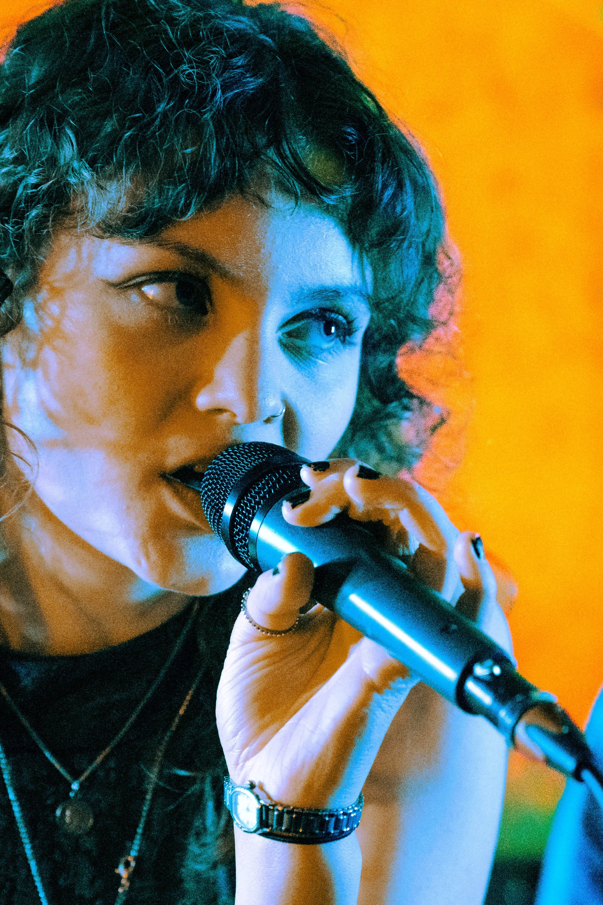 A woman singing into a microphone, with short curly hair, multiple rings and bracelets, wearing a black top, against an orange background.