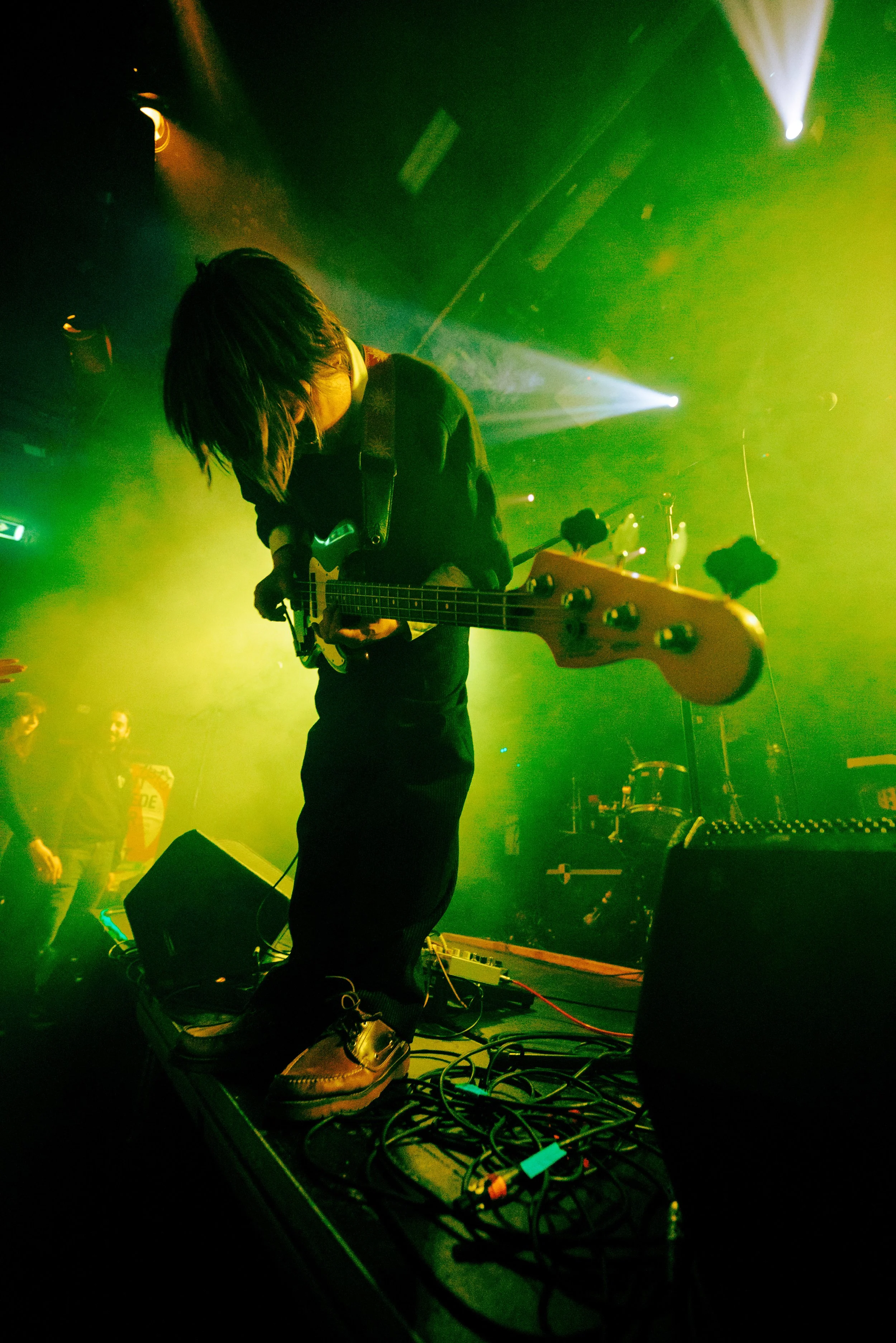 Musician performing on stage with a green spotlight, playing an electric bass guitar, with the focus on the musician's shoes and pedalboard, audience visible in the background.