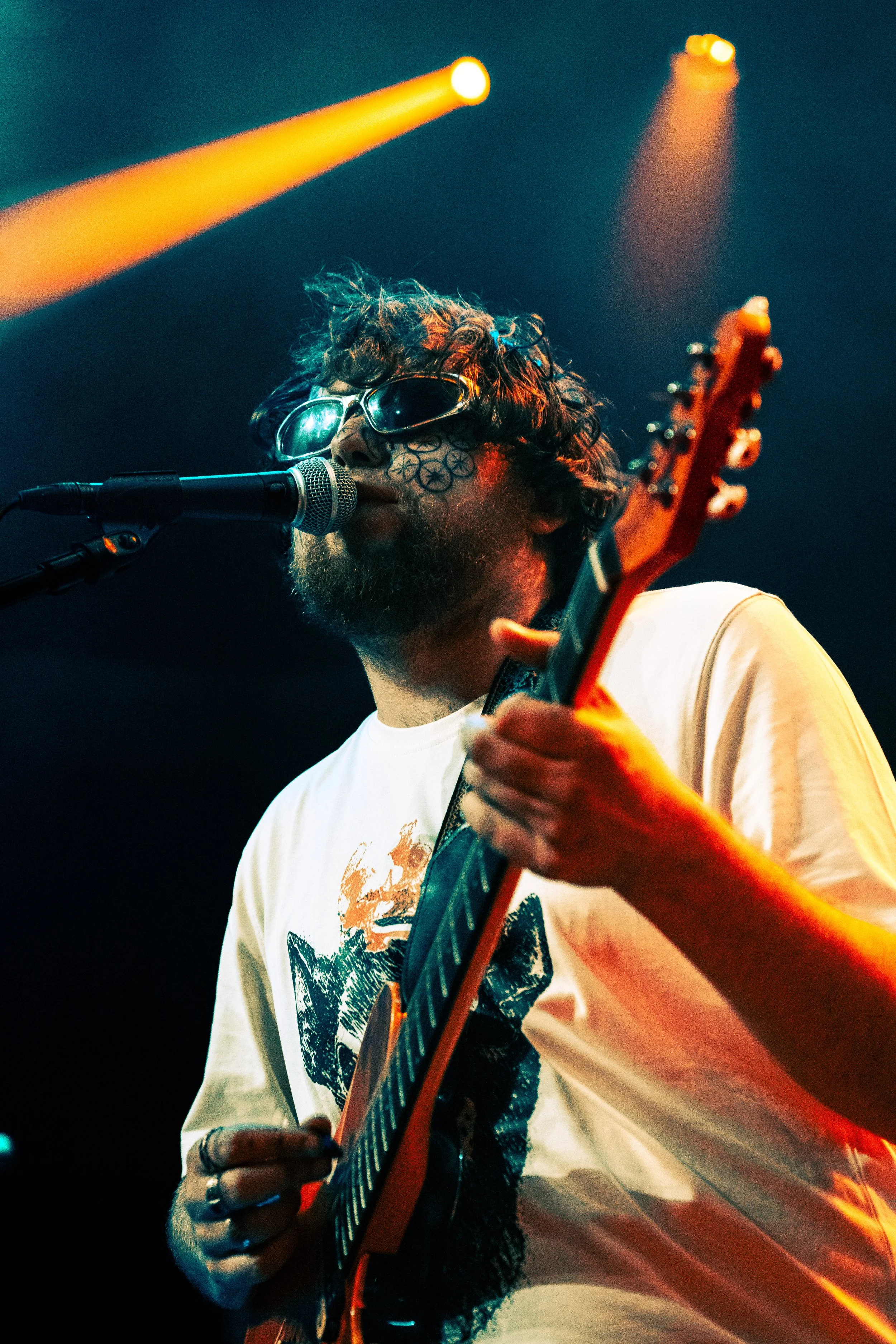 A male musician with curly hair and tattoos near his eye, wearing sunglasses, singing into a microphone while playing an electric guitar on stage with yellow stage lights overhead.