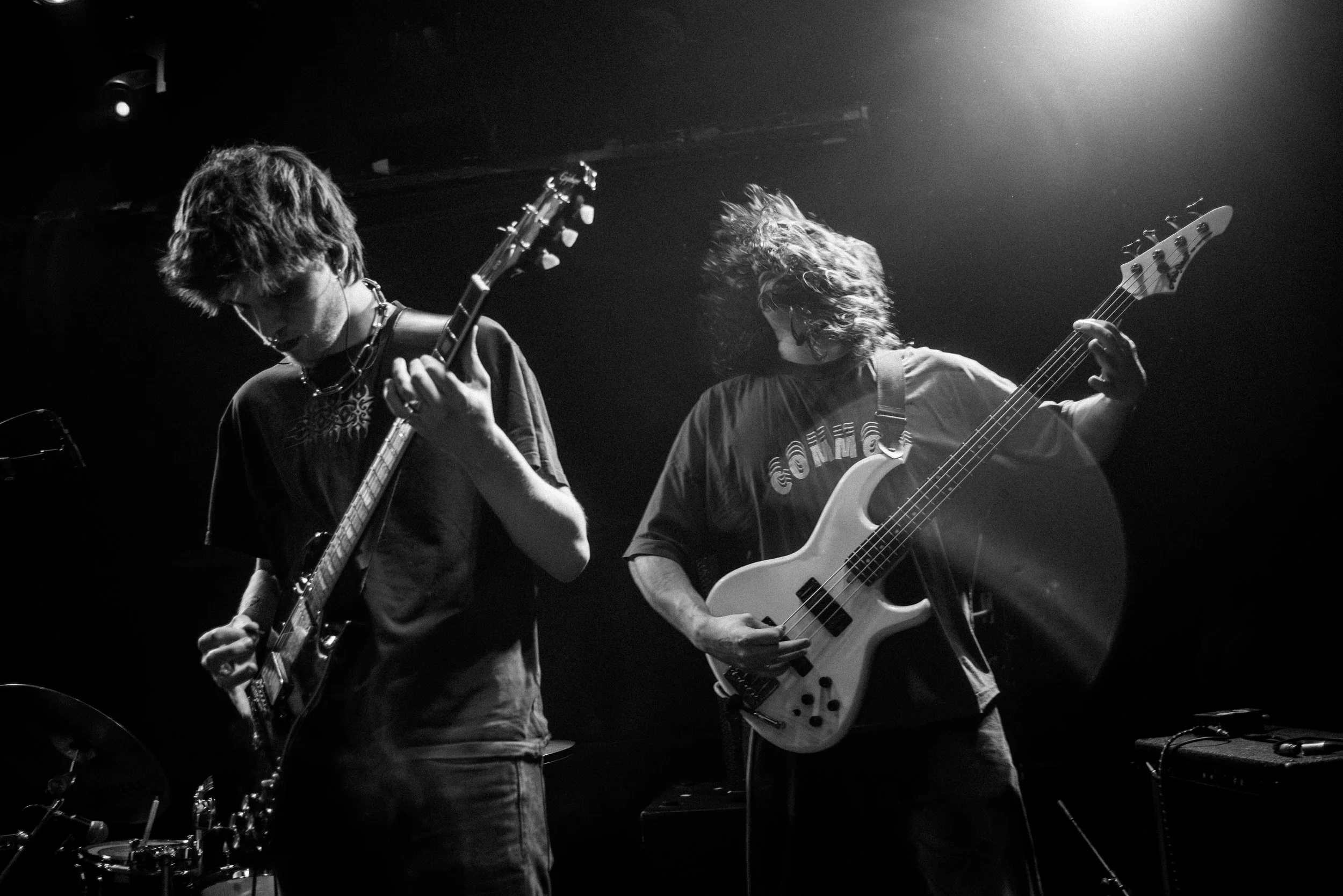 Two musicians playing electric guitar and bass on stage in a black-and-white photo, with stage lighting highlighting their movement.