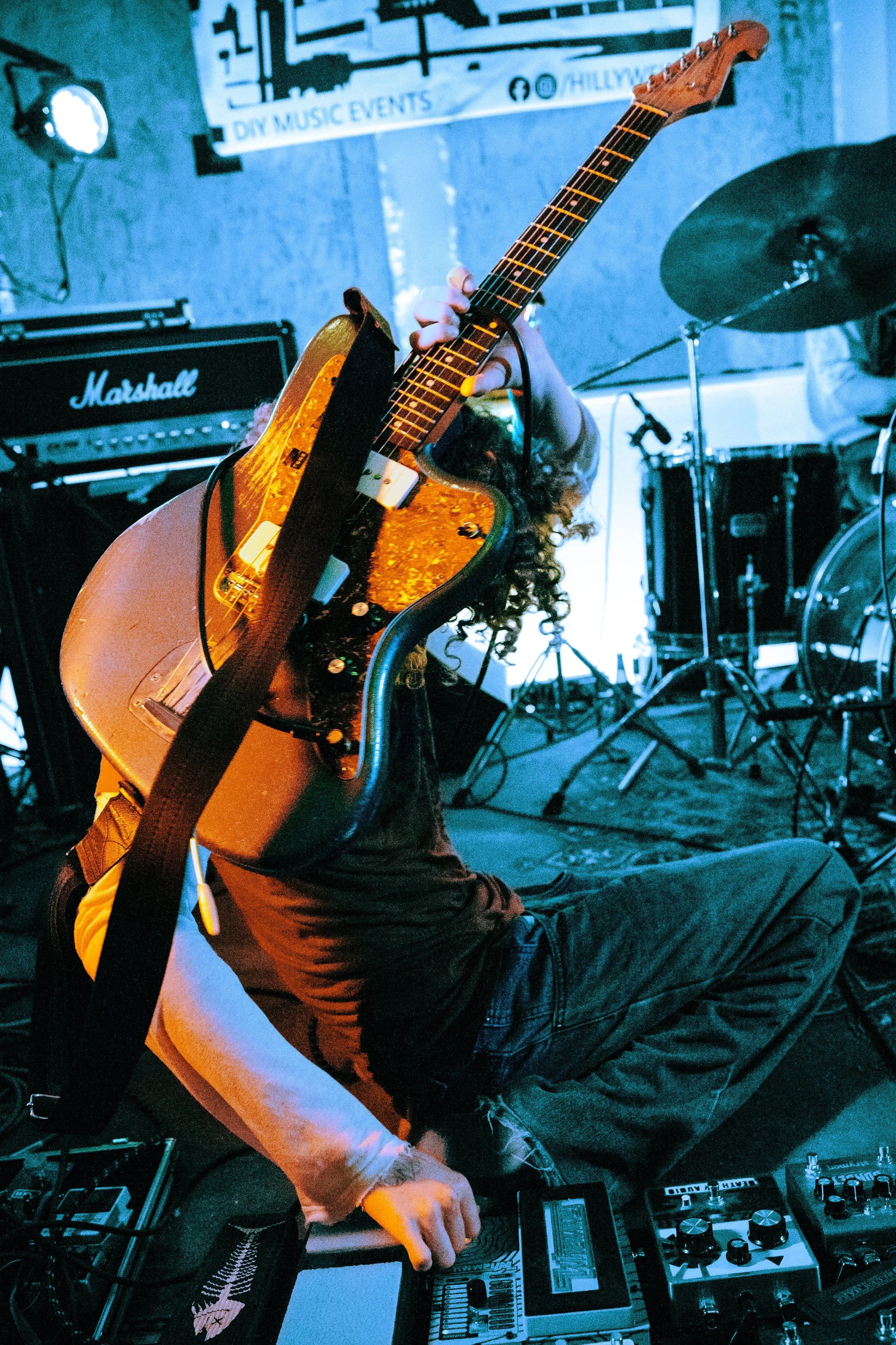 Person playing guitar and using electronic music equipment in a dimly lit space with a drum kit and Marshall amplifier visible in the background.