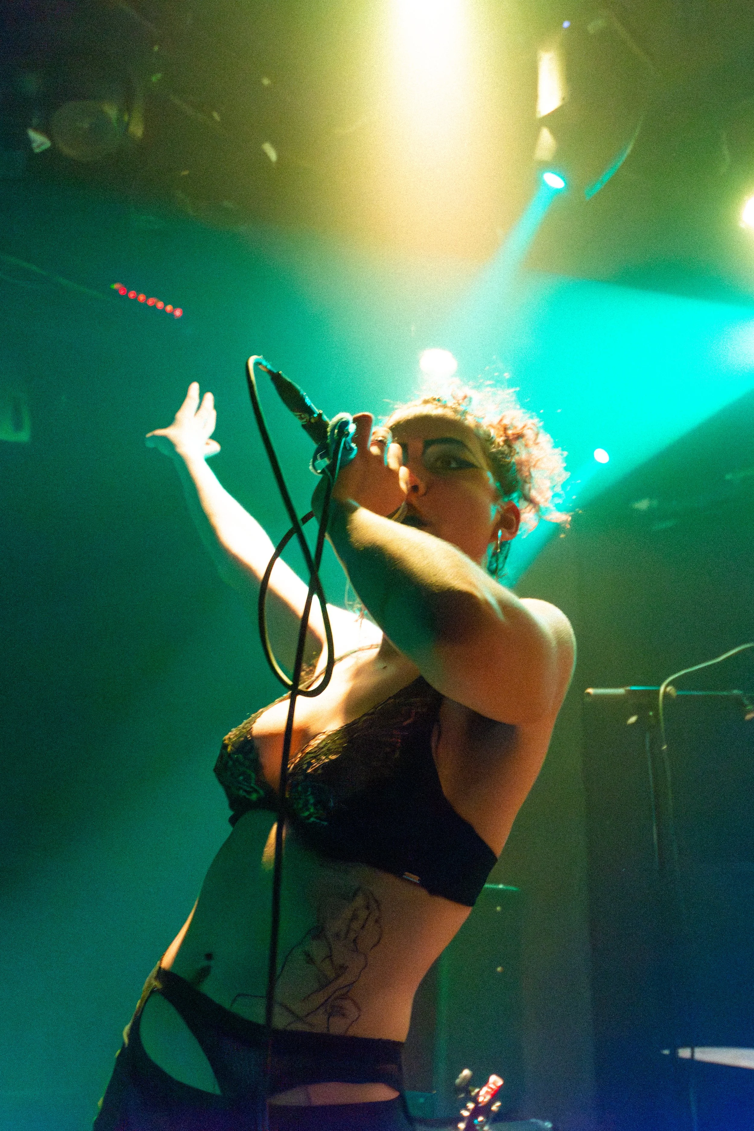 A woman with curly hair and glasses on stage, holding a microphone, with her other hand raised, wearing a black lace top and shorts, with visible tattoos on her thigh, illuminated by colored stage lights.
