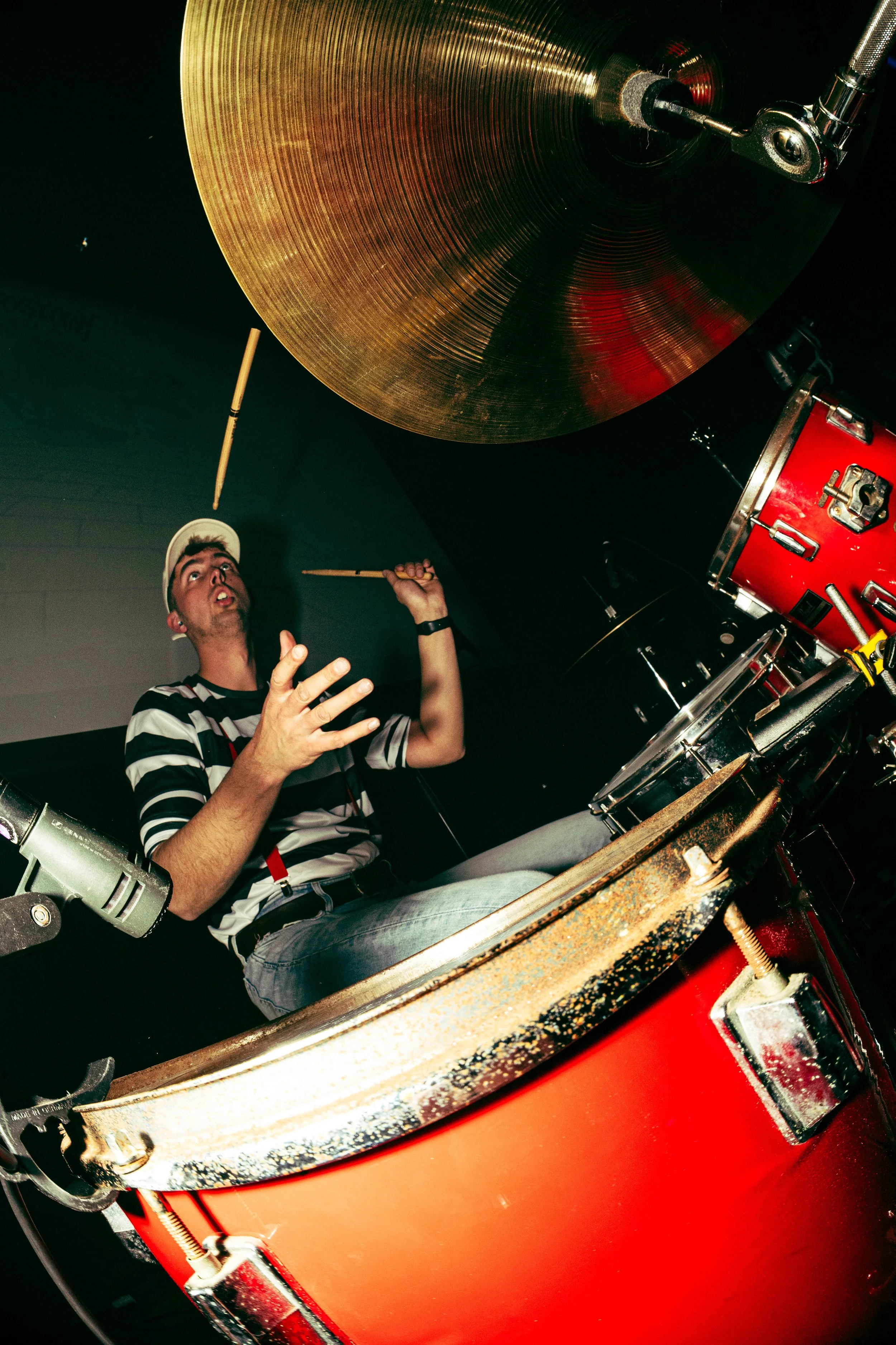 A person wearing a cap and striped shirt playing drums in a dark room, with a focus on a large, shiny cymbal and red drum set.