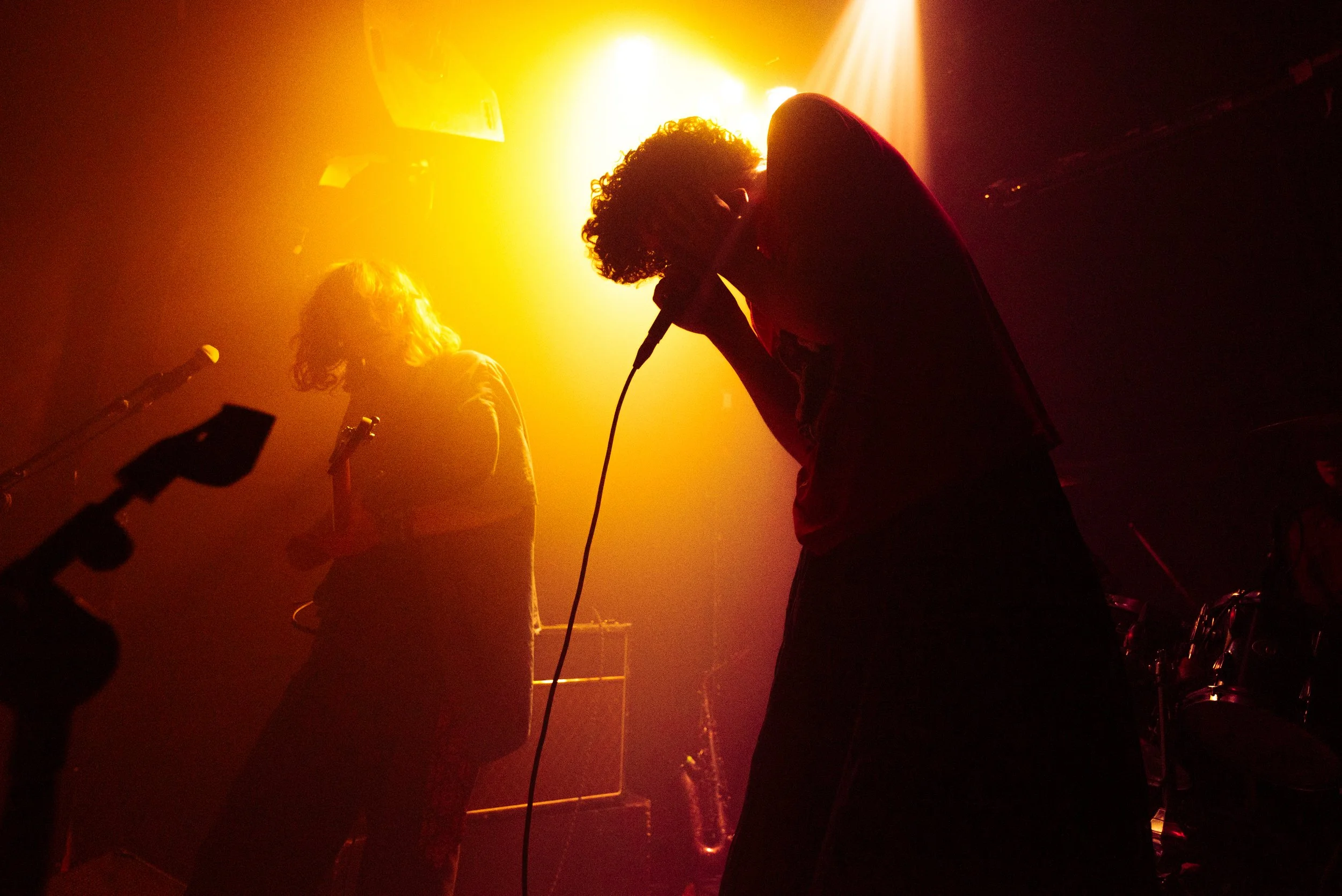 Two performers on stage, one with curly hair and the other with long hair, both holding microphones, illuminated by warm, orange-yellow stage lighting.