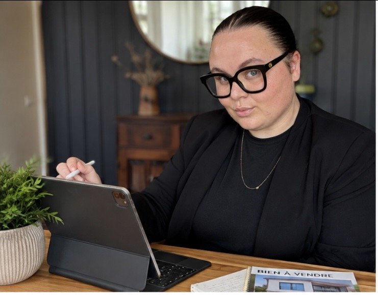 Une femme portant des lunettes noires assise à une table avec une tablette, un stylo, un livre avec la mention "BIEN À VENDRE" et une plante d'intérieur.