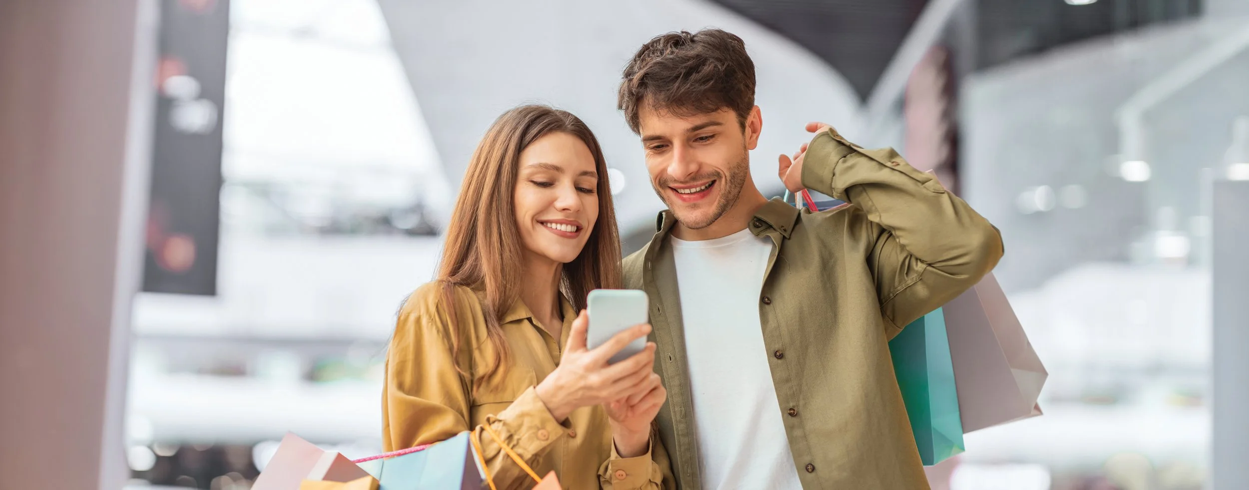A young woman and man smiling while looking at a smartphone in a shopping mall, with shopping bags in the background.