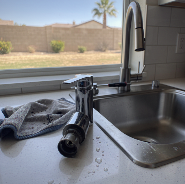 A kitchen sink with faucet and wet towel on the counter, with a backyard view through the window showing grass, fence, and pa