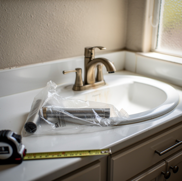 Bathroom vanity with sink, faucet, and toilet seat next to a window with a plastic-wrapped pipe and measuring tape on the cou