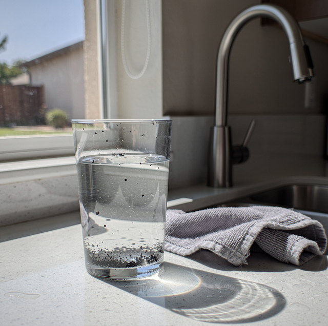 Glass of water on a kitchen countertop next to a faucet and towel by a window.
