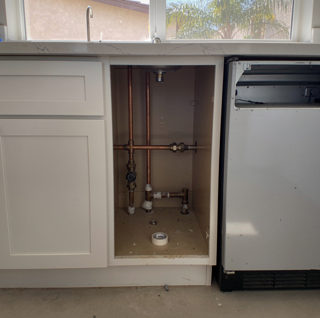 Open cabinet under kitchen counter showing plumbing pipes and fittings next to a dishwasher.
