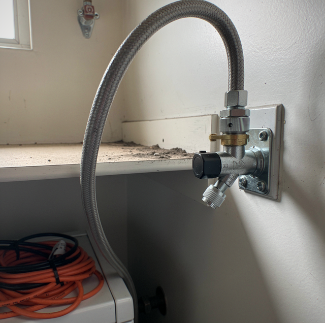 A braided metal hose connects to the side of a white toilet via a valve in a bathroom.