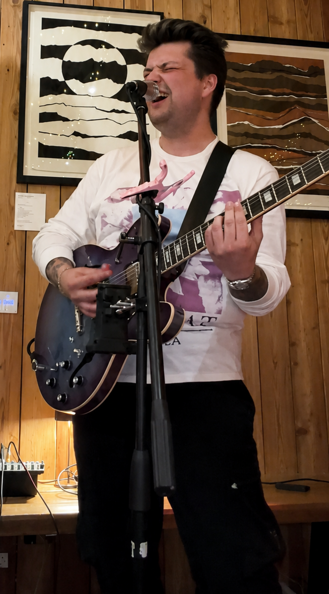 A man is singing passionately into a microphone while playing an electric guitar in a wooden-paneled room with abstract art on the wall behind him.